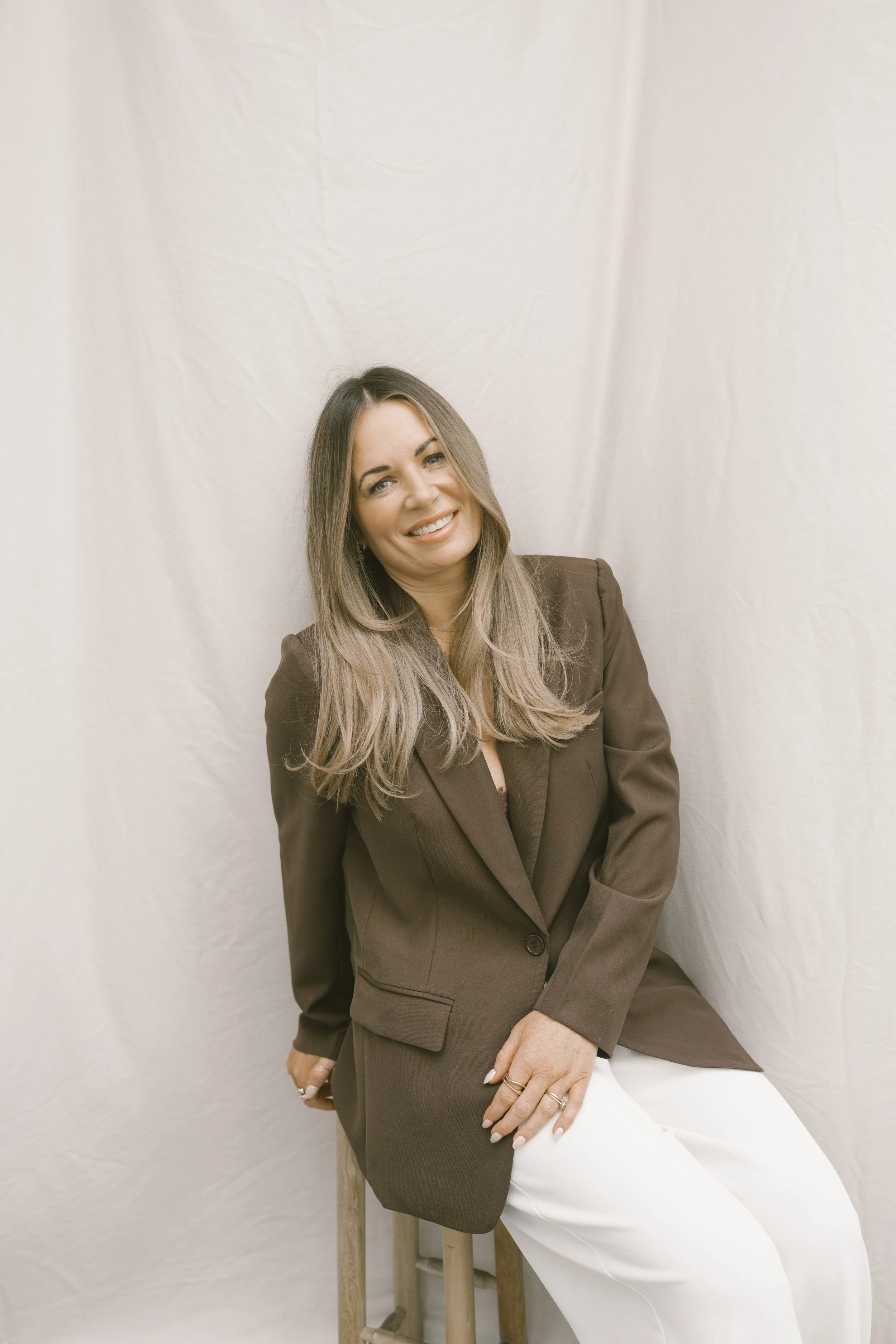 A woman with long wiavy blonde hair, wearing a brown blazer and white pants, sitting on a stool against a white backdrop, smiling at the camera.