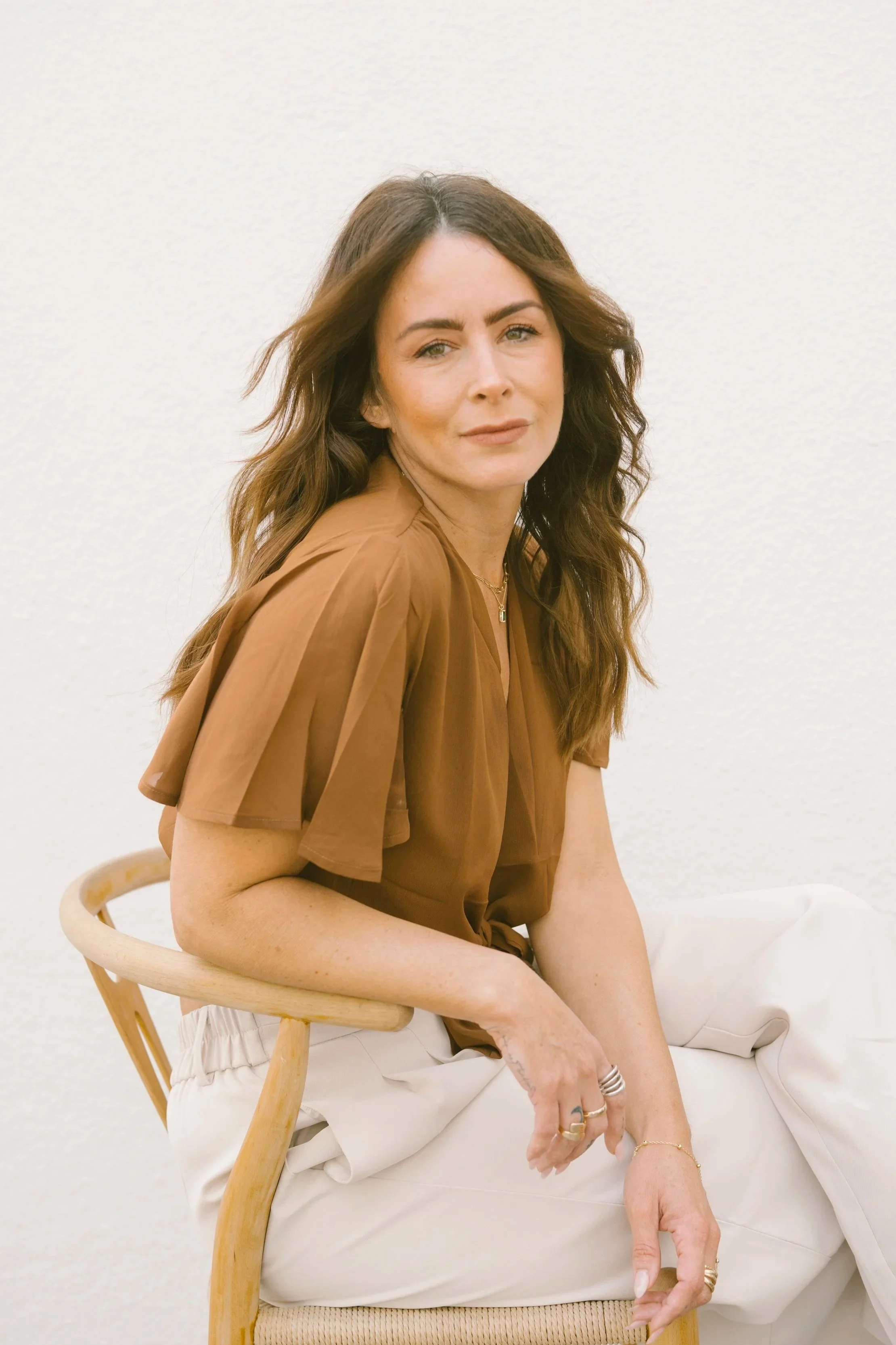 A woman with long brown hair wearing a light brown blouse and white pants, seated on a wooden chair against a plain white wall.