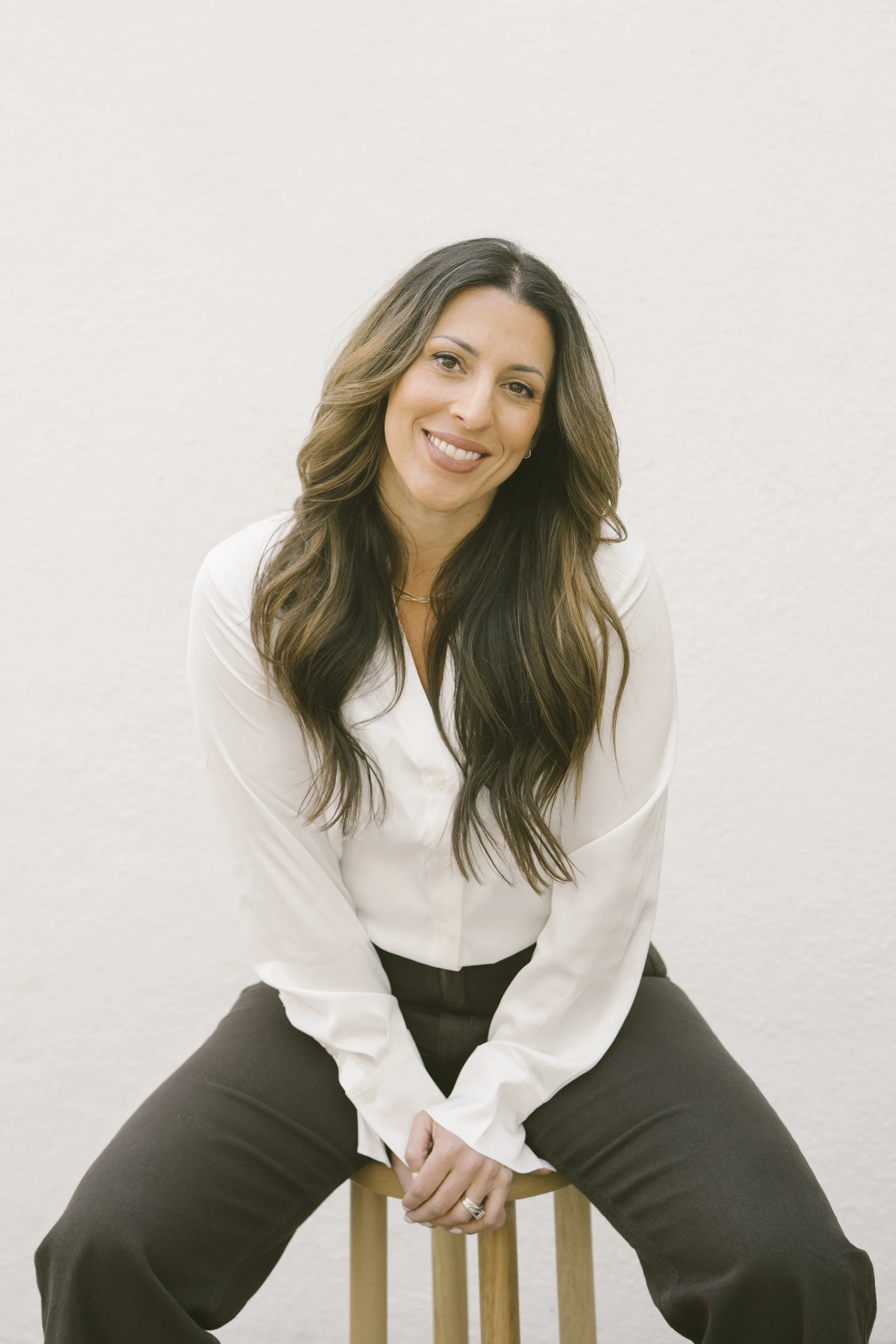 A woman with long, wavy brown hair and a warm smile, sitting on a wooden stool against a plain white wall, wearing a white blouse and dark pants.