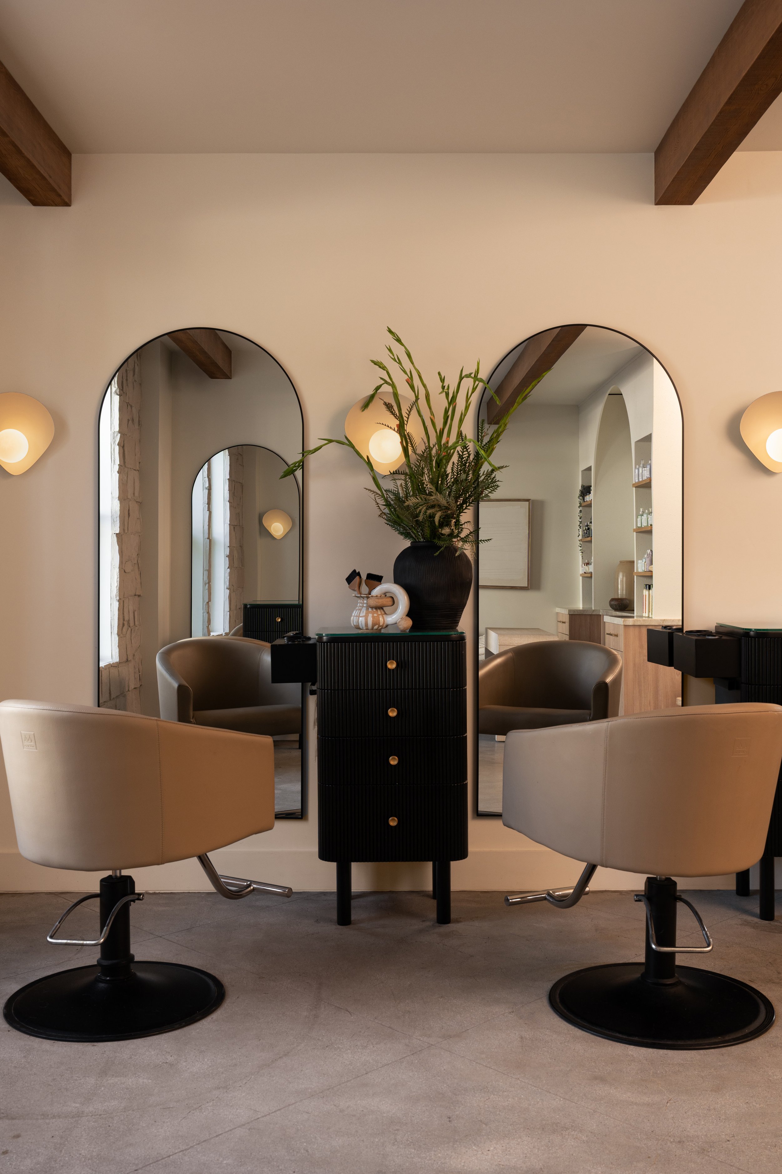 A modern interior with two black salon chairs, a black cabinet with a large vase holding green plants, and three mirrors on the wall. The space is decorated with warm lighting and has wooden beams on the ceiling.