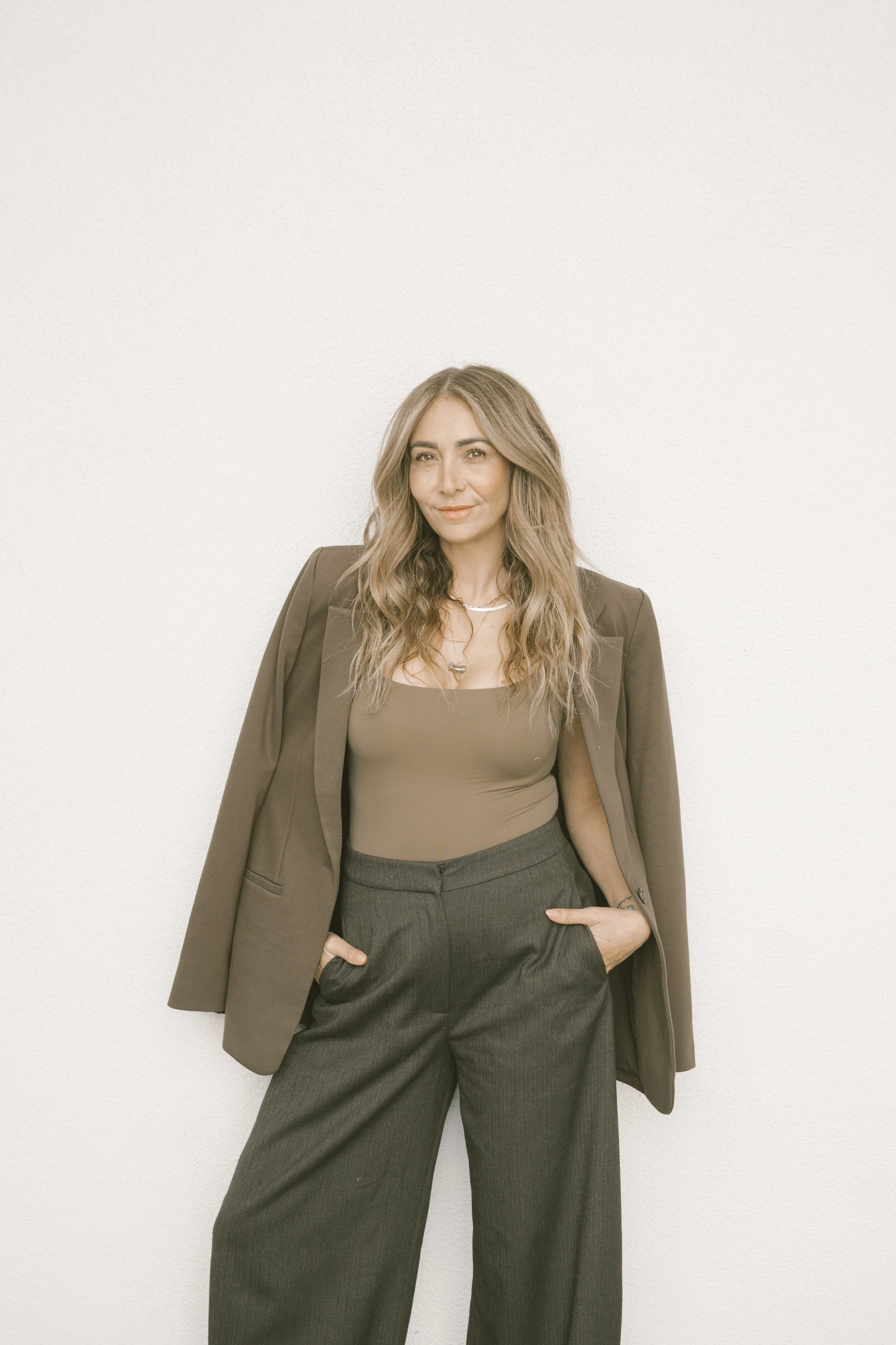 A woman with wavy light brown hair wearing a brown blazer over a taupe top, black high-waisted pleated pants, and accessorized with necklaces, standing against a plain white wall, with one hand in her pocket.