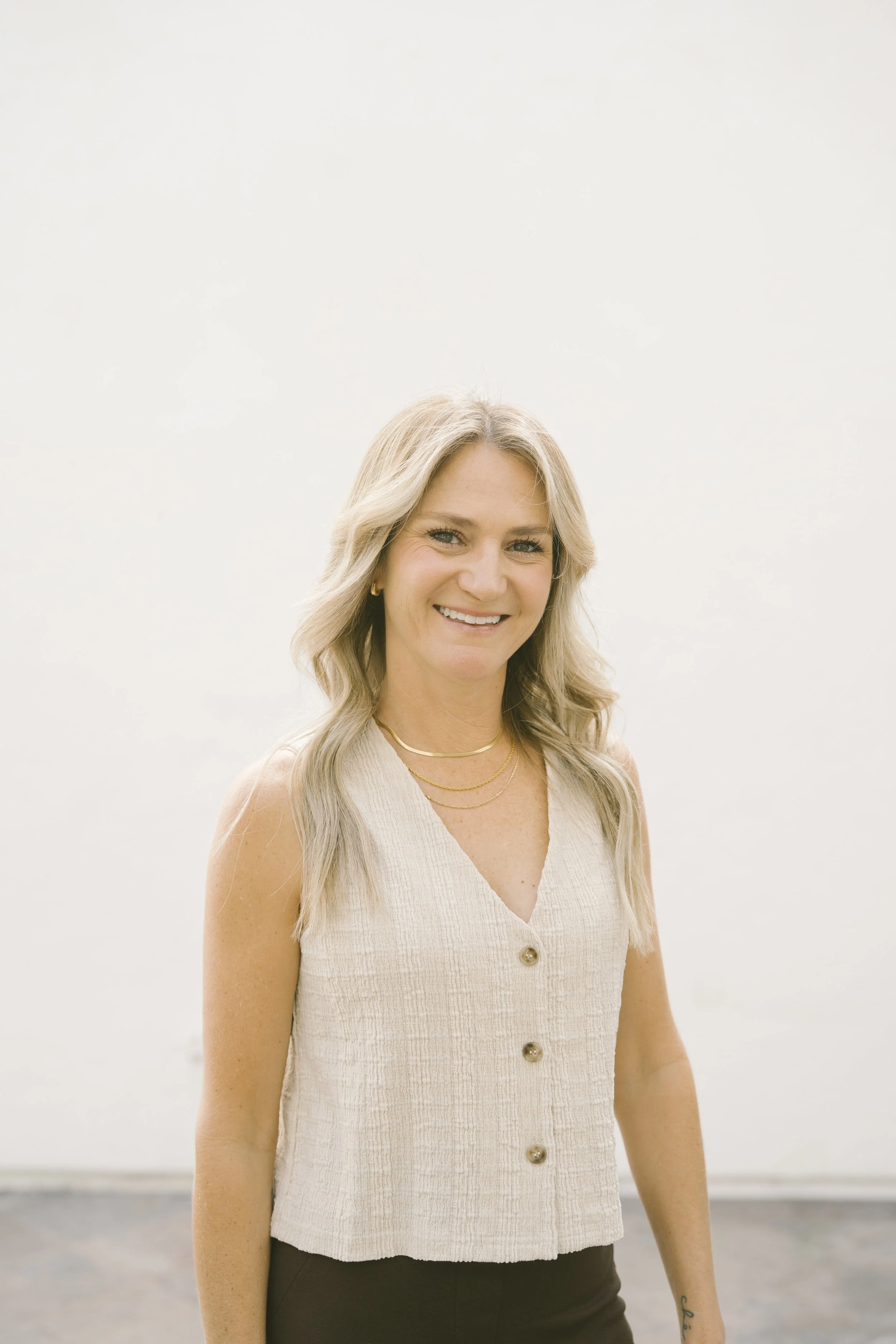 Close-up of a smiling woman with long blonde hair, wearing a light sleeveless button-up top and layered gold necklaces, standing outdoors against a light, neutral background.