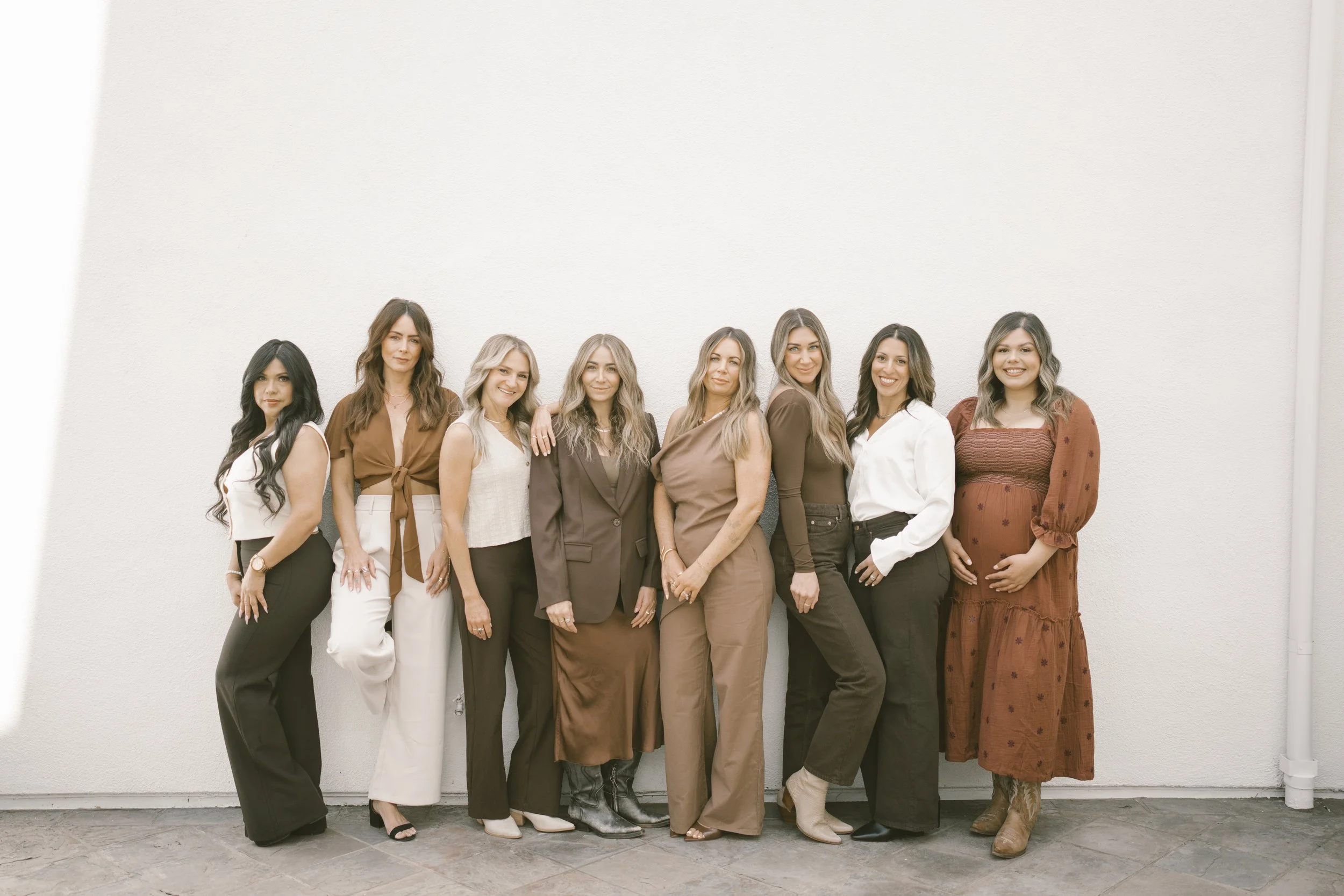 A group of nine women standing side by side against a plain white wall, all smiling and dressed in stylish casual and professional clothing.