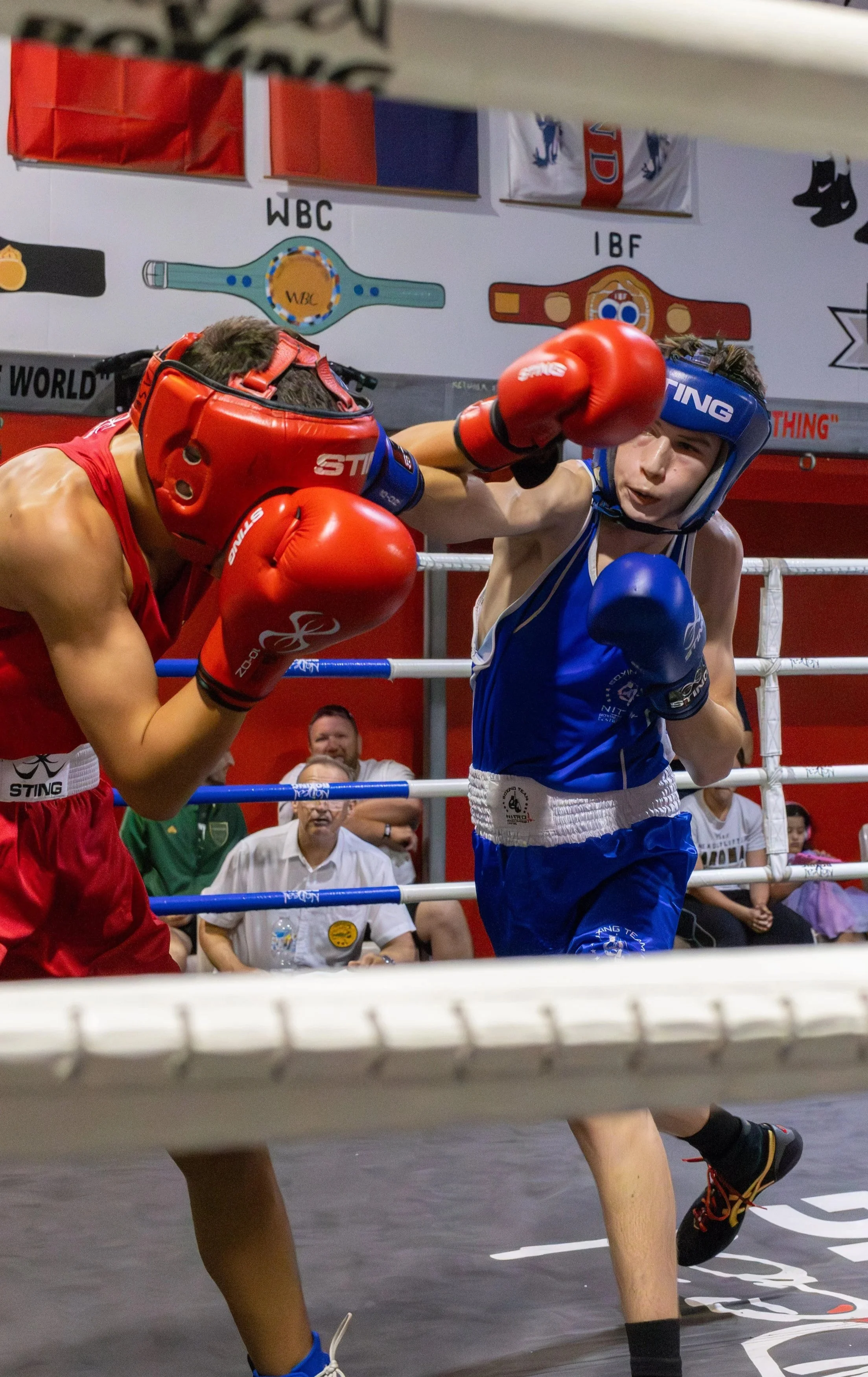 Two amateur boxers in a match, wearing protective headgear and gloves, engaged in close combat inside a boxing ring with a referee and spectators in the background.