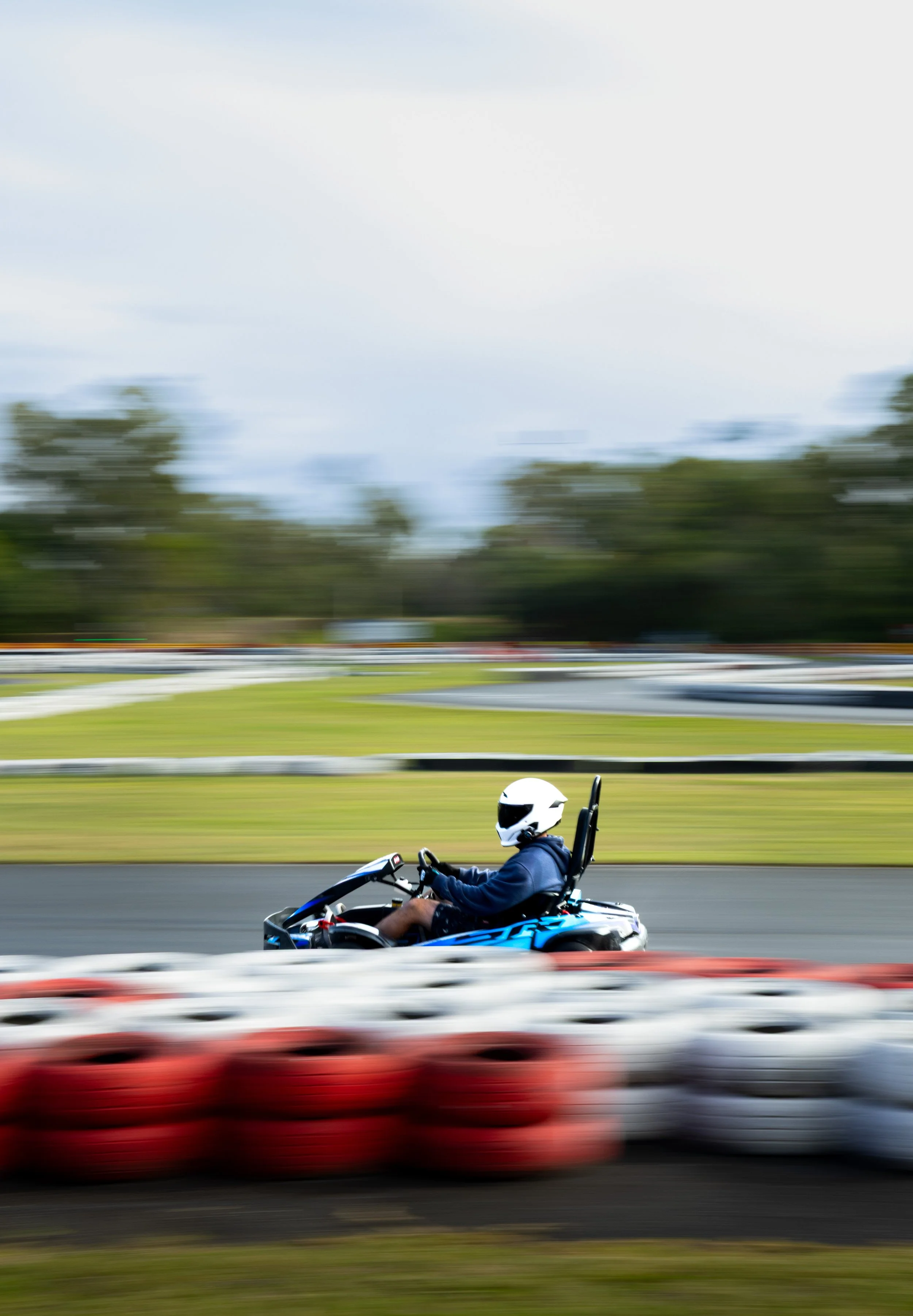 A man riding a go-kart on a race track, wearing a white helmet and a dark jacket, with blurred red cars passing by in the foreground and trees in the background.