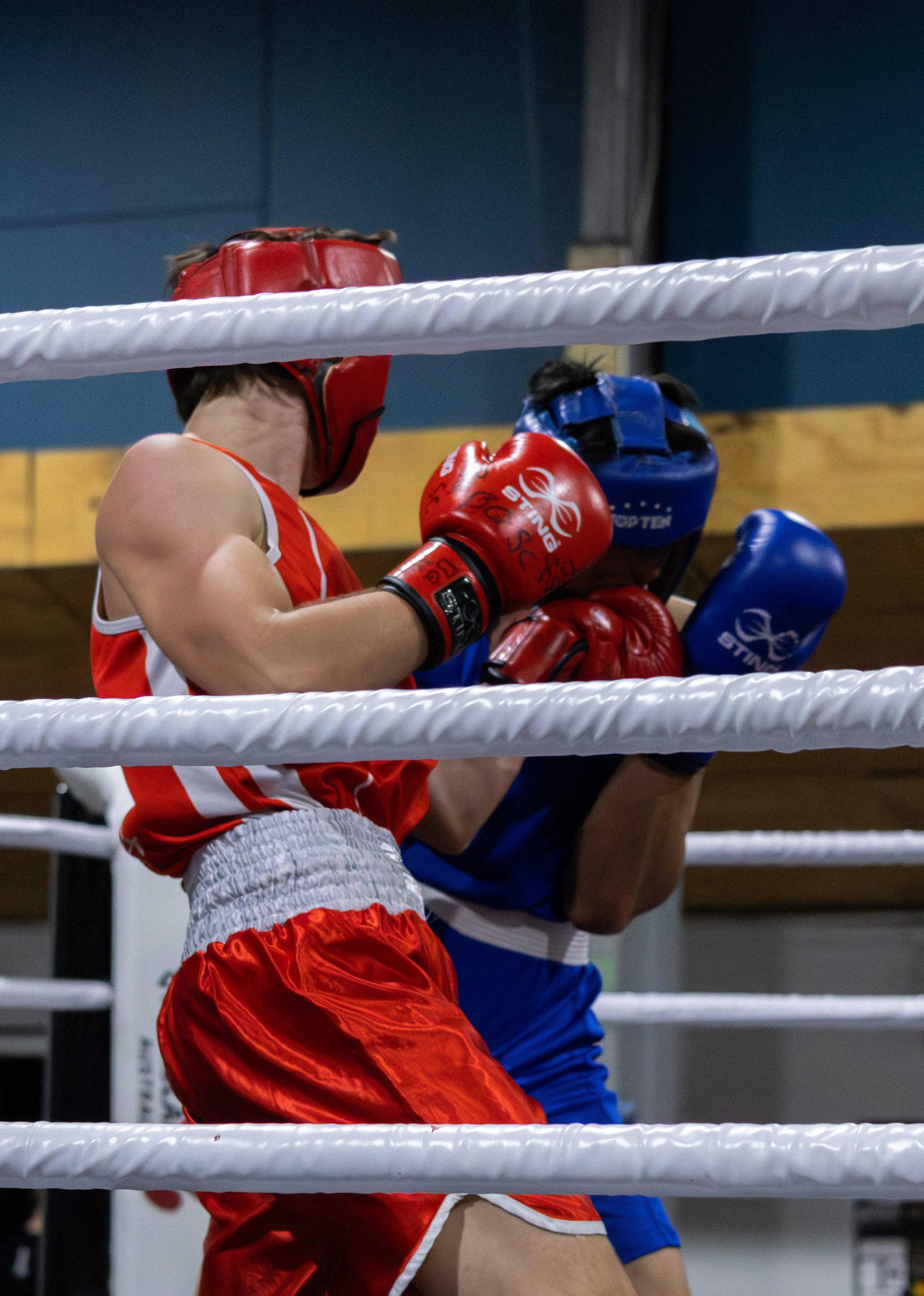 Two young male boxers wearing headgear and gloves fighting in a boxing ring, with one in red and the other in blue.