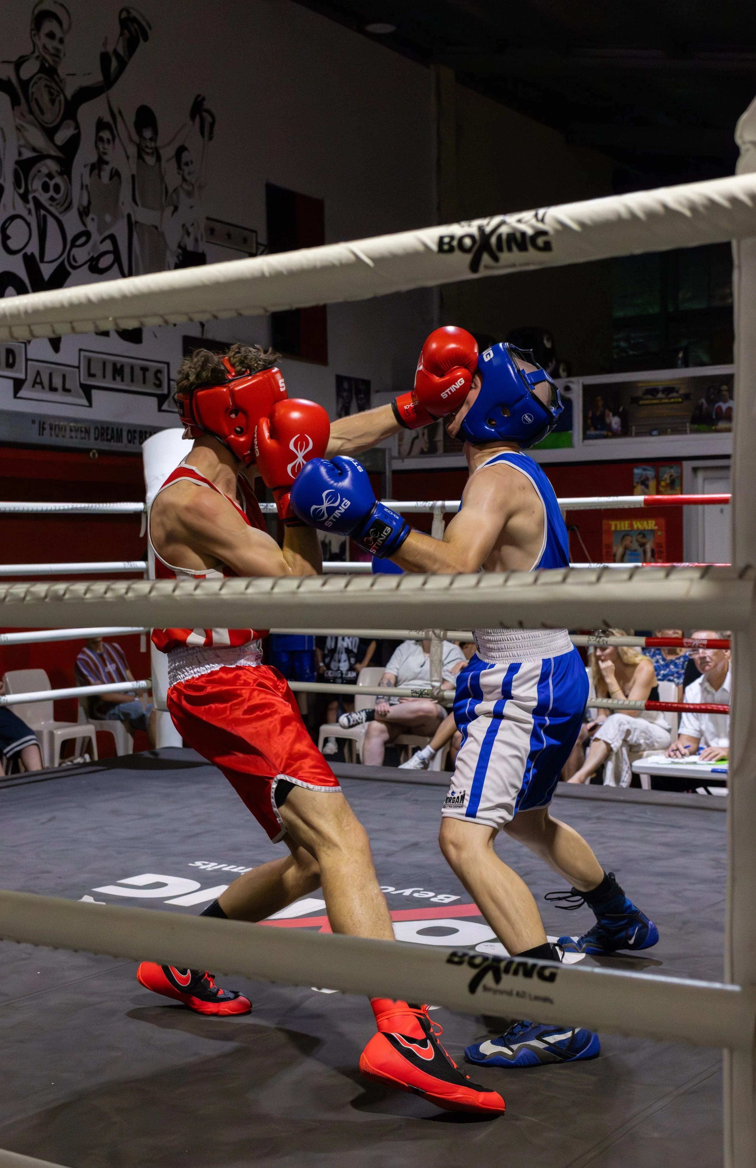Two boxers in a boxing ring engaged in a match. One is wearing red gloves, red shorts, and red shoes, while the other is wearing blue gloves, blue and white shorts, and blue shoes. The boxer in red is throwing a punch at the boxer in blue, who is blocking.