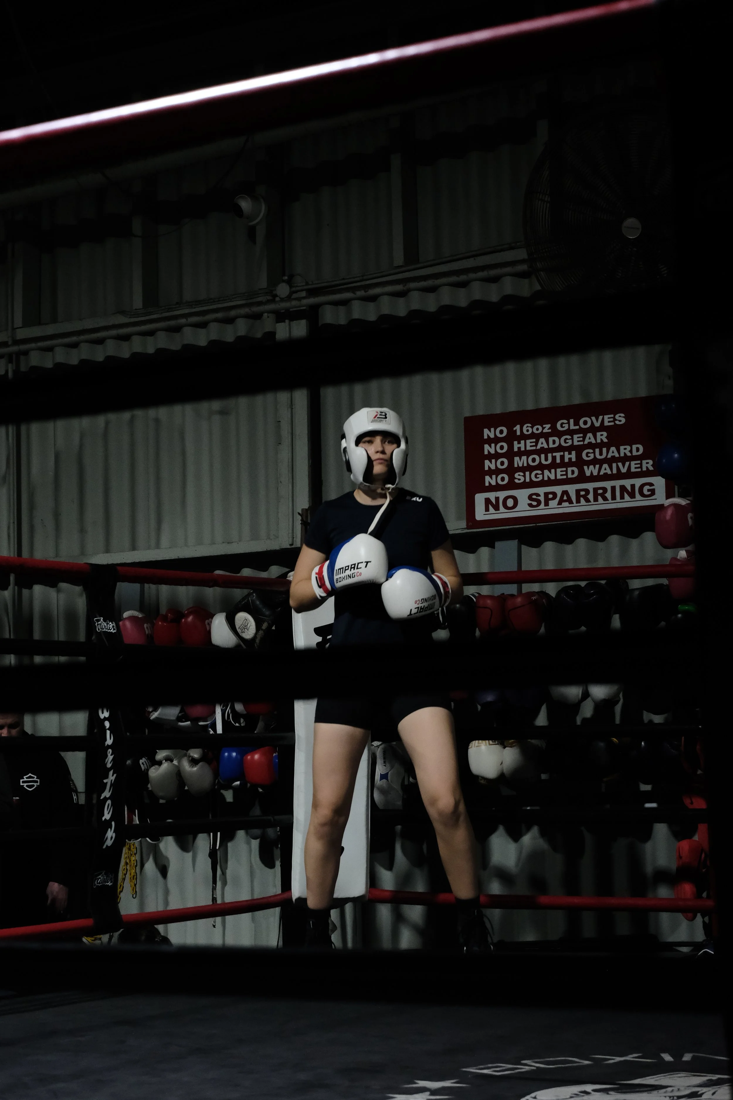 A woman stands inside a boxing ring, wearing boxing gloves, a white headgear, and athletic clothing, with a serious expression. Behind her, boxing gear and a sign with rules are visible.