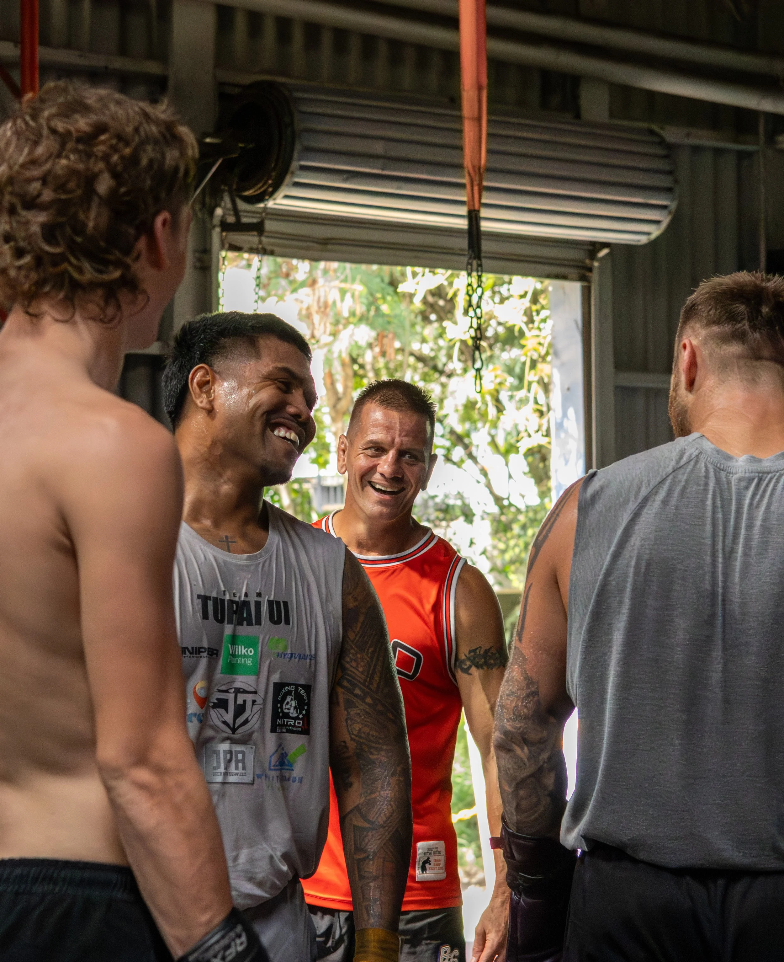 Group of shirtless male athletes smiling and talking in a gym or locker room, with some wearing athletic shorts and tattoos visible on their arms and necks.