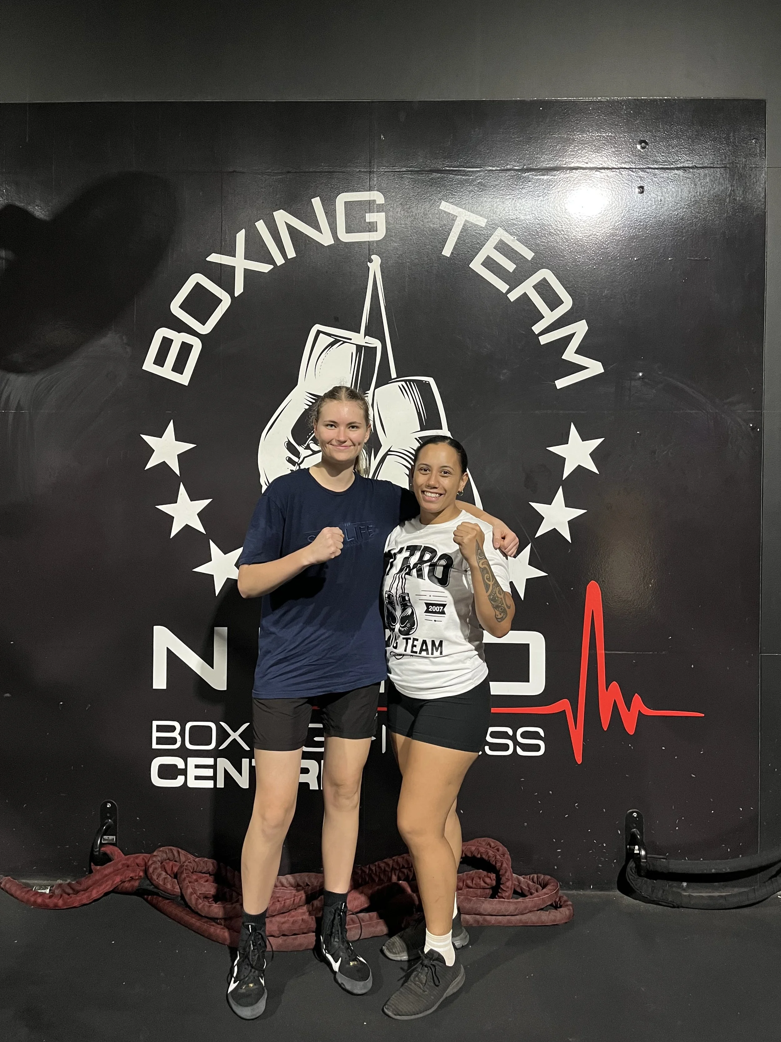 Two women standing in front of a black fitness center wall that reads 'Boxing Team' and features boxing gloves and stars. The woman on the left has blonde hair, wears a blue T-shirt, black shorts, and boxing shoes. The woman on the right has dark hair, tattoos on her arms, wears a white T-shirt with a boxing graphic, black shorts, and black sneakers. Both women are posing with clenched fists.
