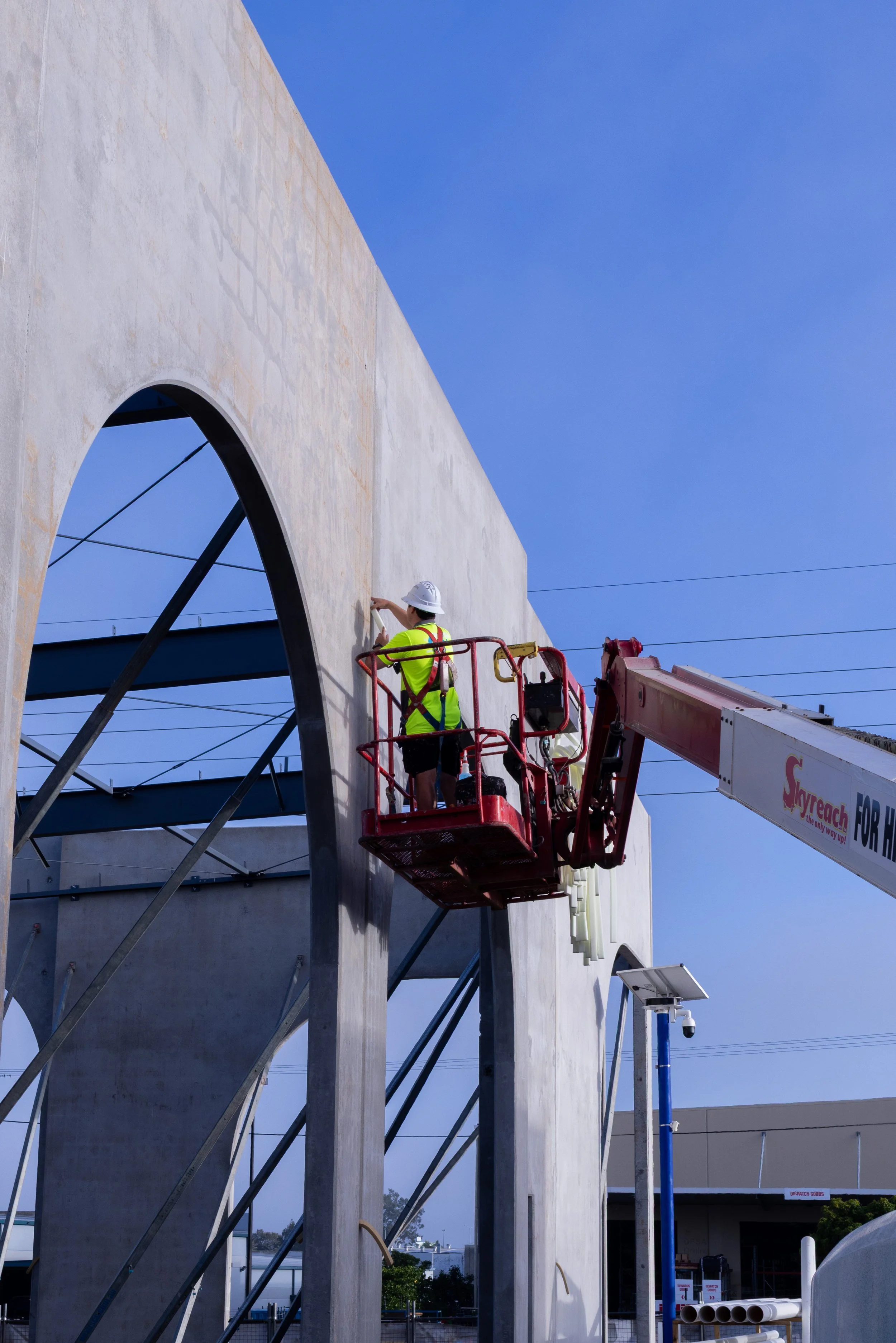 Construction worker on a cherry picker installing a large concrete structure with an arched opening, under bright blue sky.