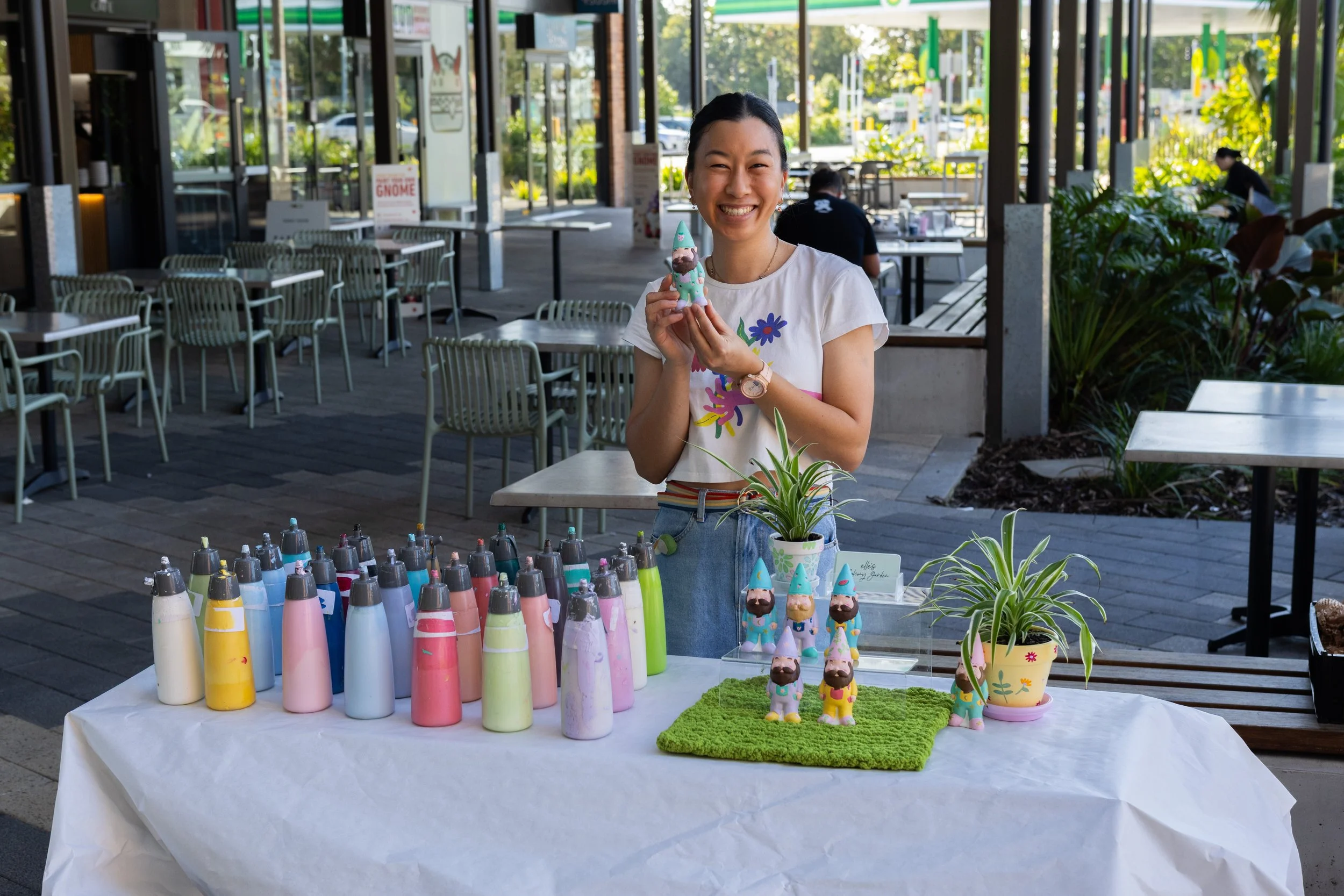 A woman smiling and holding a decorated gnome figurine at a craft or art market stand outside with colorful bottles of paint, gnome figurines, potted plants, and decorative items on a table. The background shows a glass building with tables and people inside.