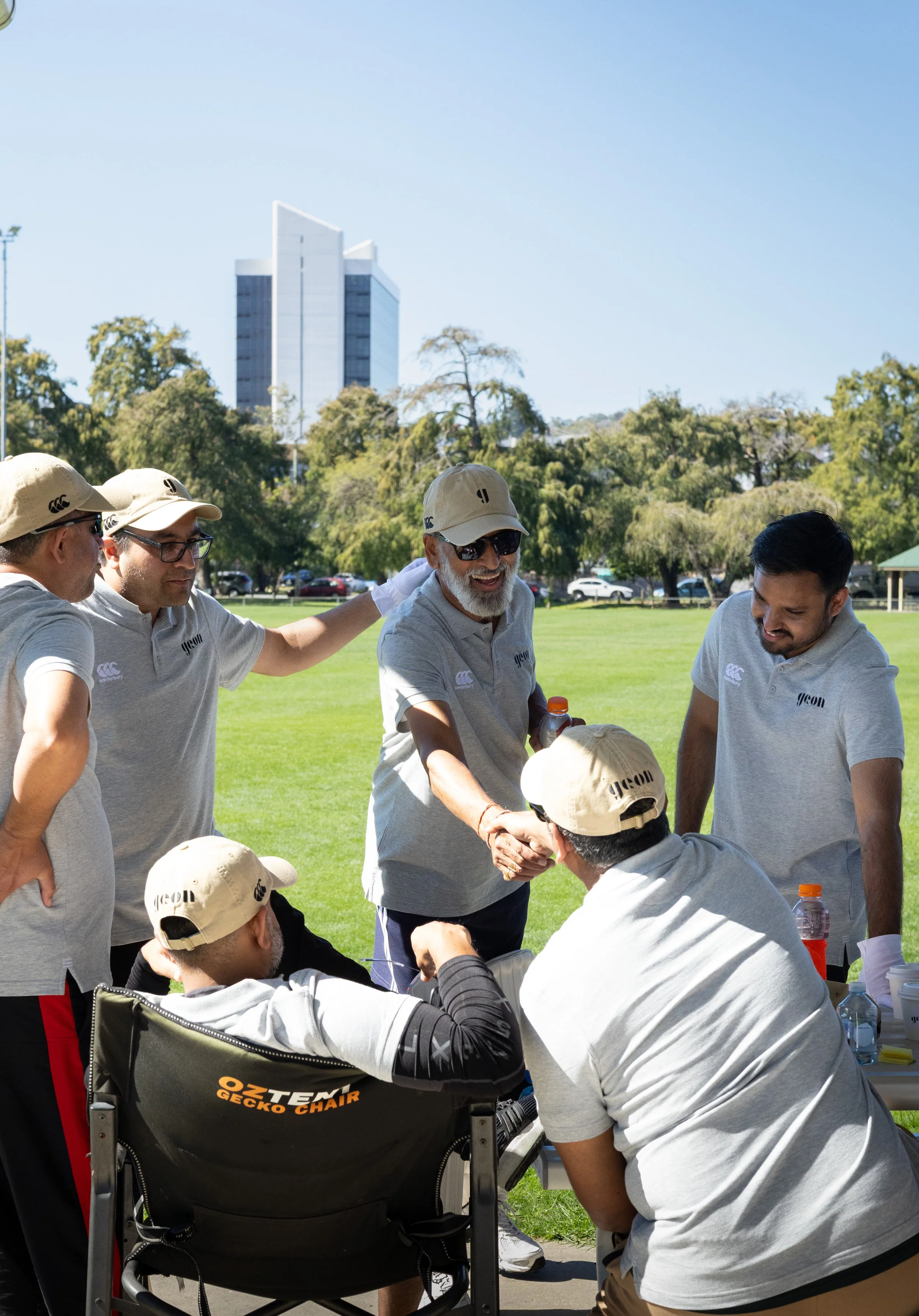 Group of people in gray shirts and beige caps gathered outdoors on a sunny day, some smiling and shaking hands, with trees, a grassy field, and a cityscape in the background.