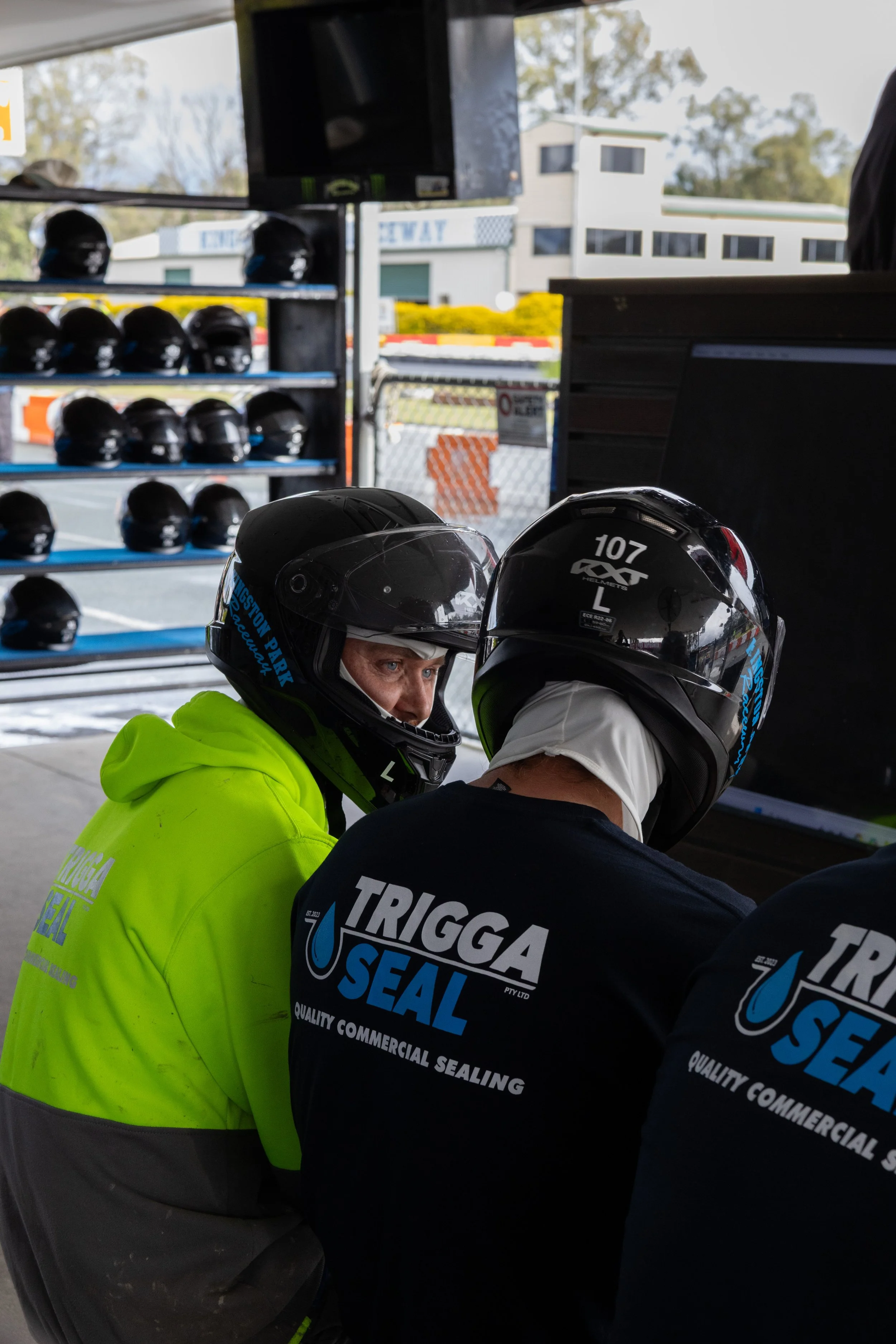 Two people wearing motorcycle helmets and black shirts with 'TRIGGA SEAL' logo appear to be working on a task inside a garage or workshop. One person is wearing a bright yellow high-visibility jacket.