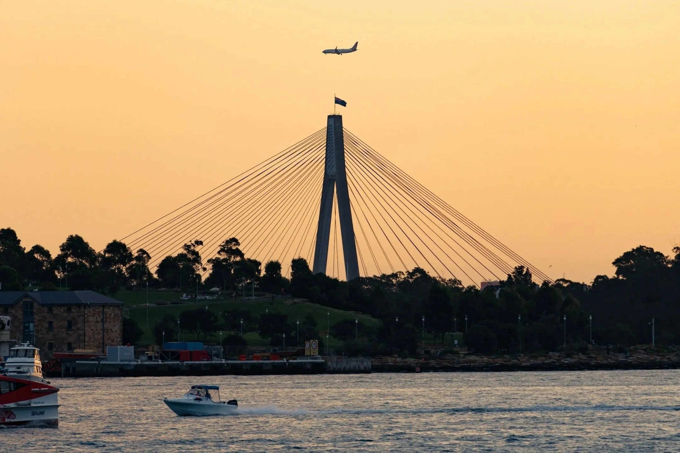 View of a river with boats, a bridge with cables and a flag, and an airplane flying in the orange sky at sunset.