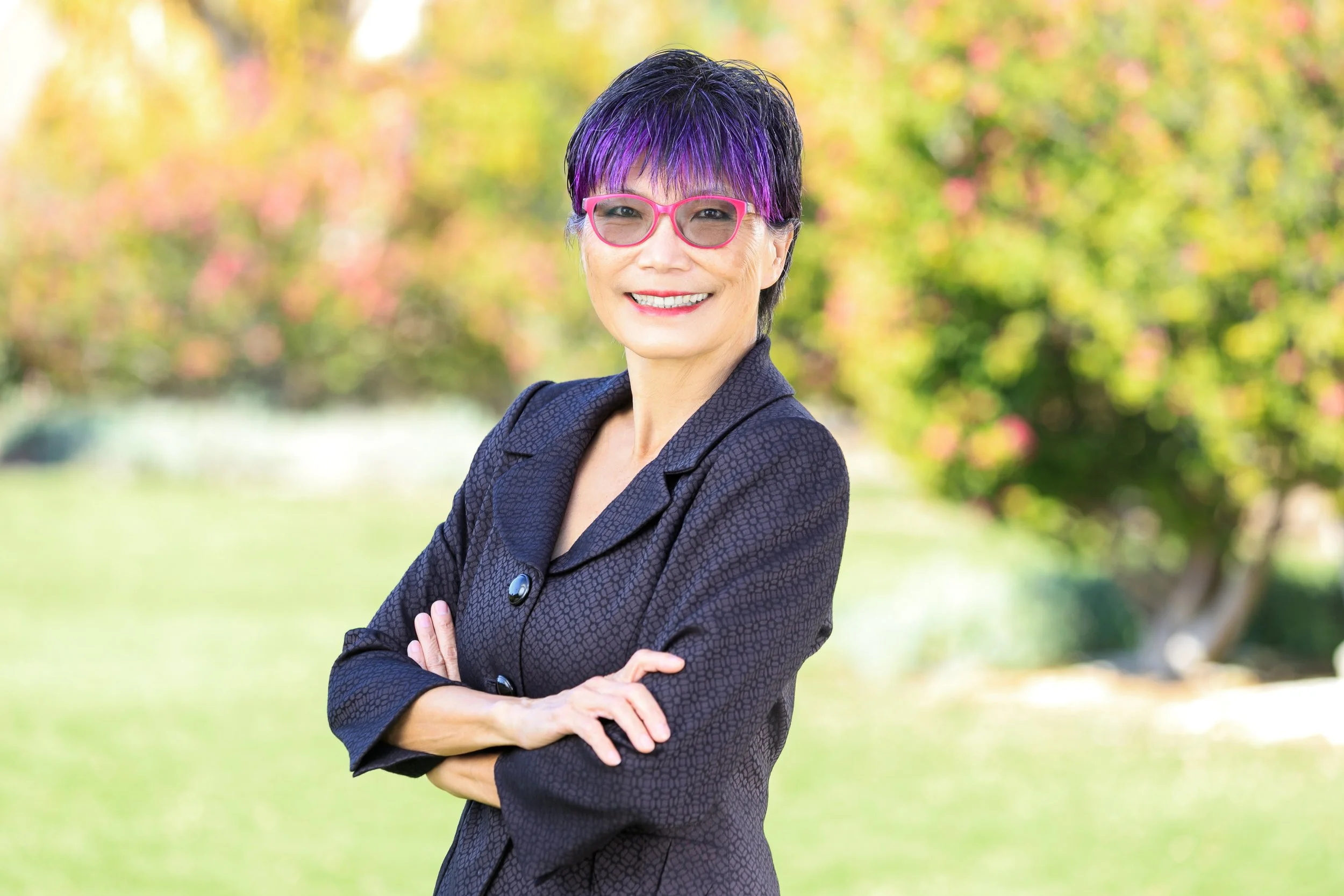 A smiling woman with short, purple-highlighted black hair wearing pink glasses and a dark patterned blazer standing outdoors with arms crossed, blurred colorful trees in the background.