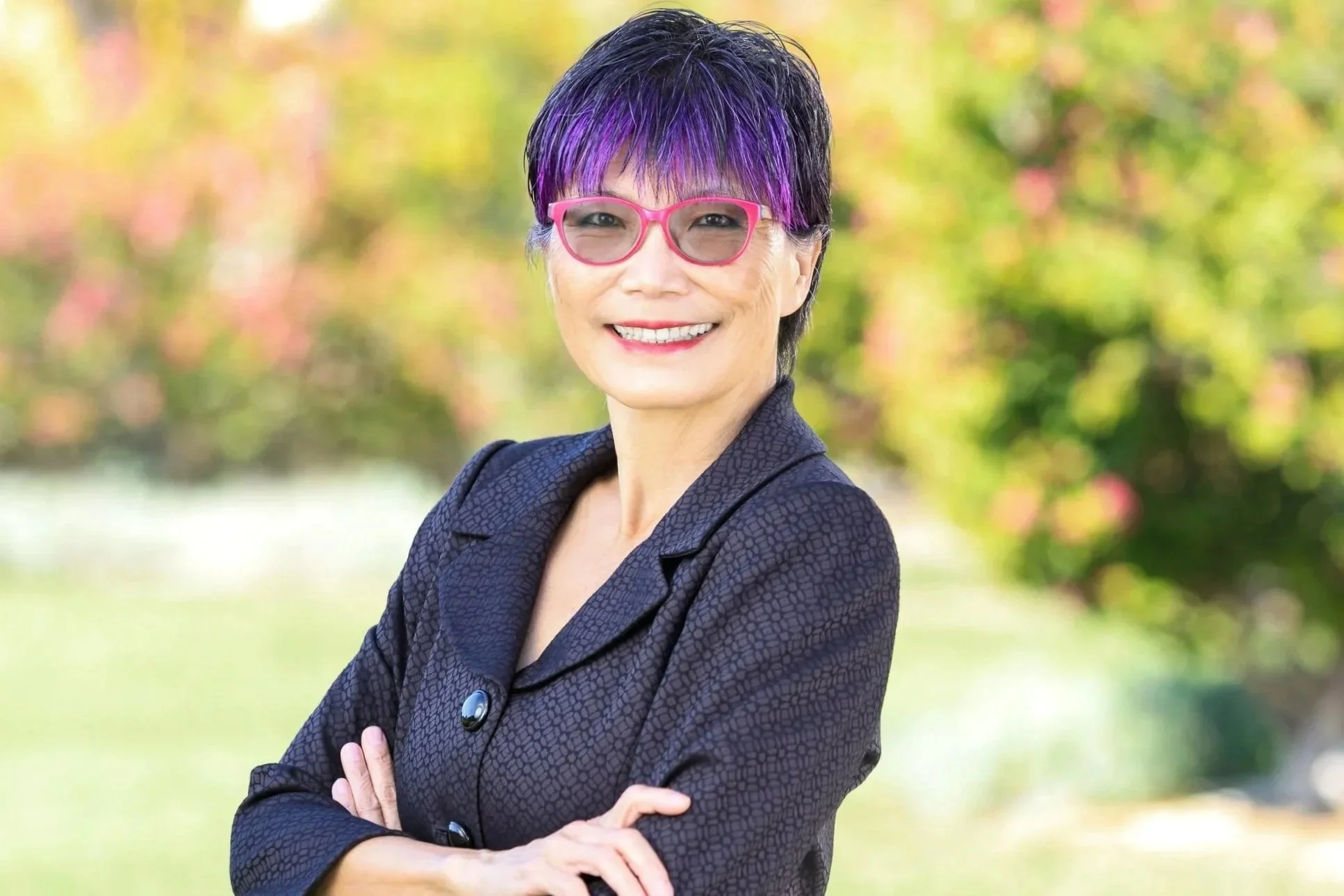 A woman with short black hair with purple highlights, wearing pink glasses and a dark patterned blazer, smiling outdoors with blurred colorful foliage in the background.