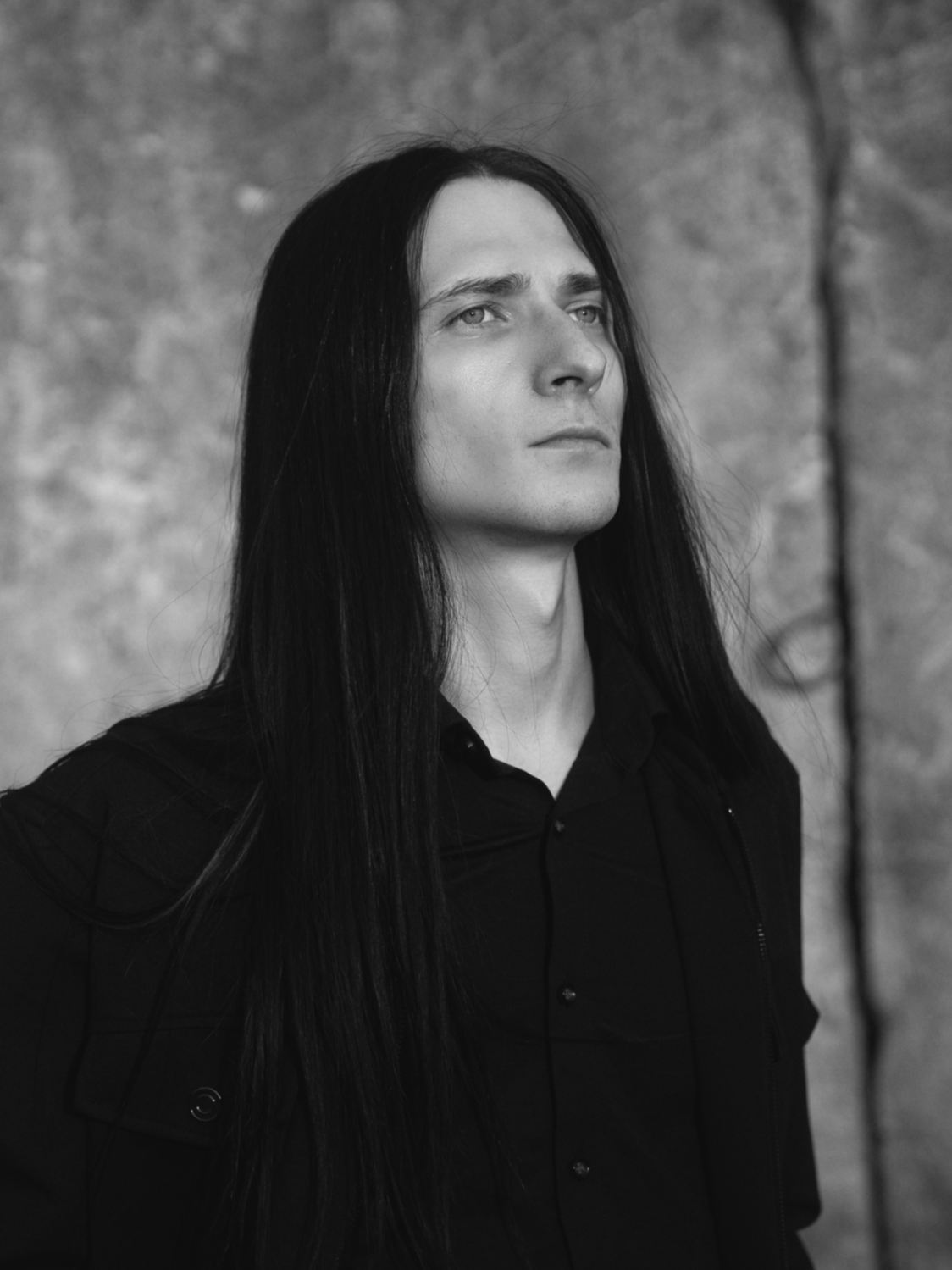 A man with long black hair wearing a black shirt and jacket, looking slightly to the side, with a blurred natural background.