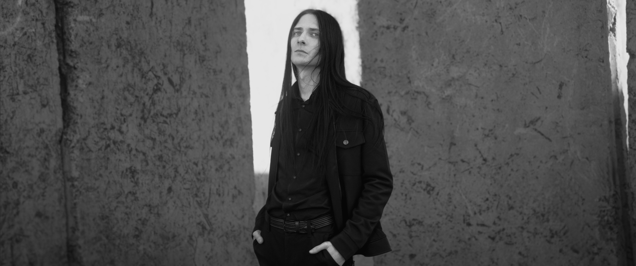 A woman with long dark hair wearing a black jacket and shirt, standing between concrete walls, looking serious.
