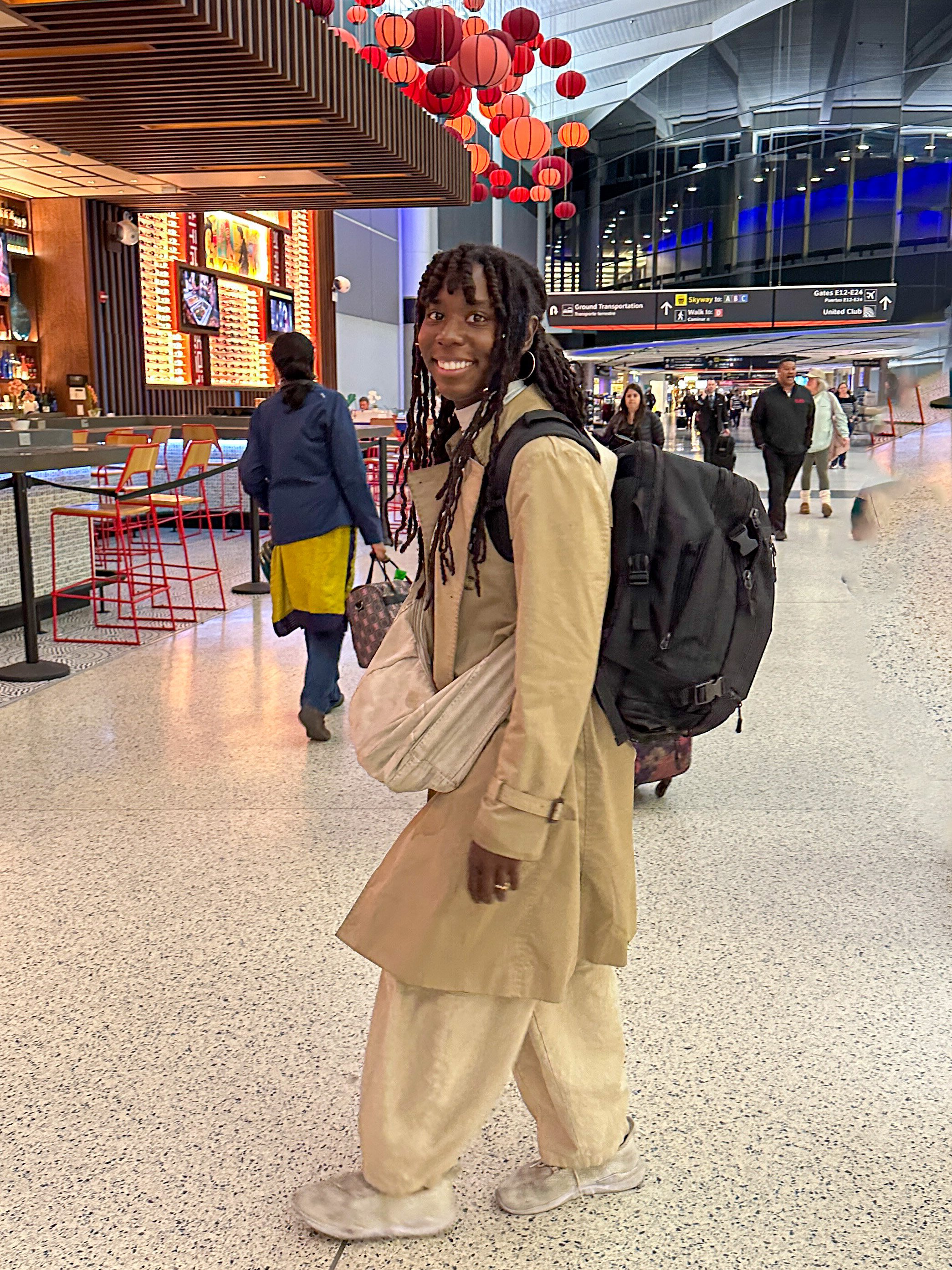 A young woman with long twists hairstyle and hoop earrings is smiling at the camera in an airport terminal. She is wearing a beige trench coat, baggy beige pants, and sneakers, and has a backpack on her back. The background shows other travelers, a bar, and decorative red lanterns hanging from the ceiling.