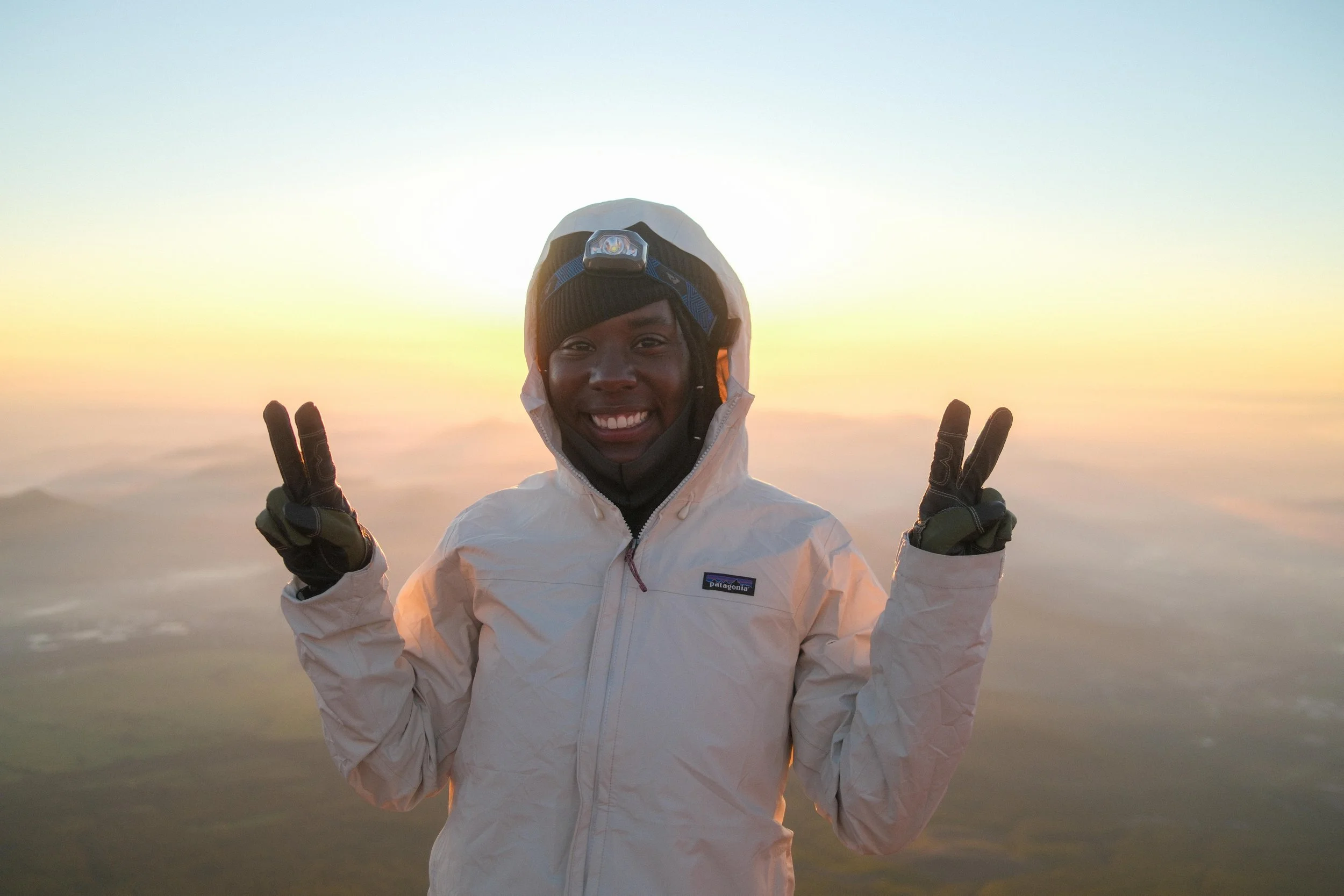 A smiling person wearing a white Patagonia jacket, a beanie, and a headlamp, standing outdoors at sunset or sunrise, making peace signs with both hands.