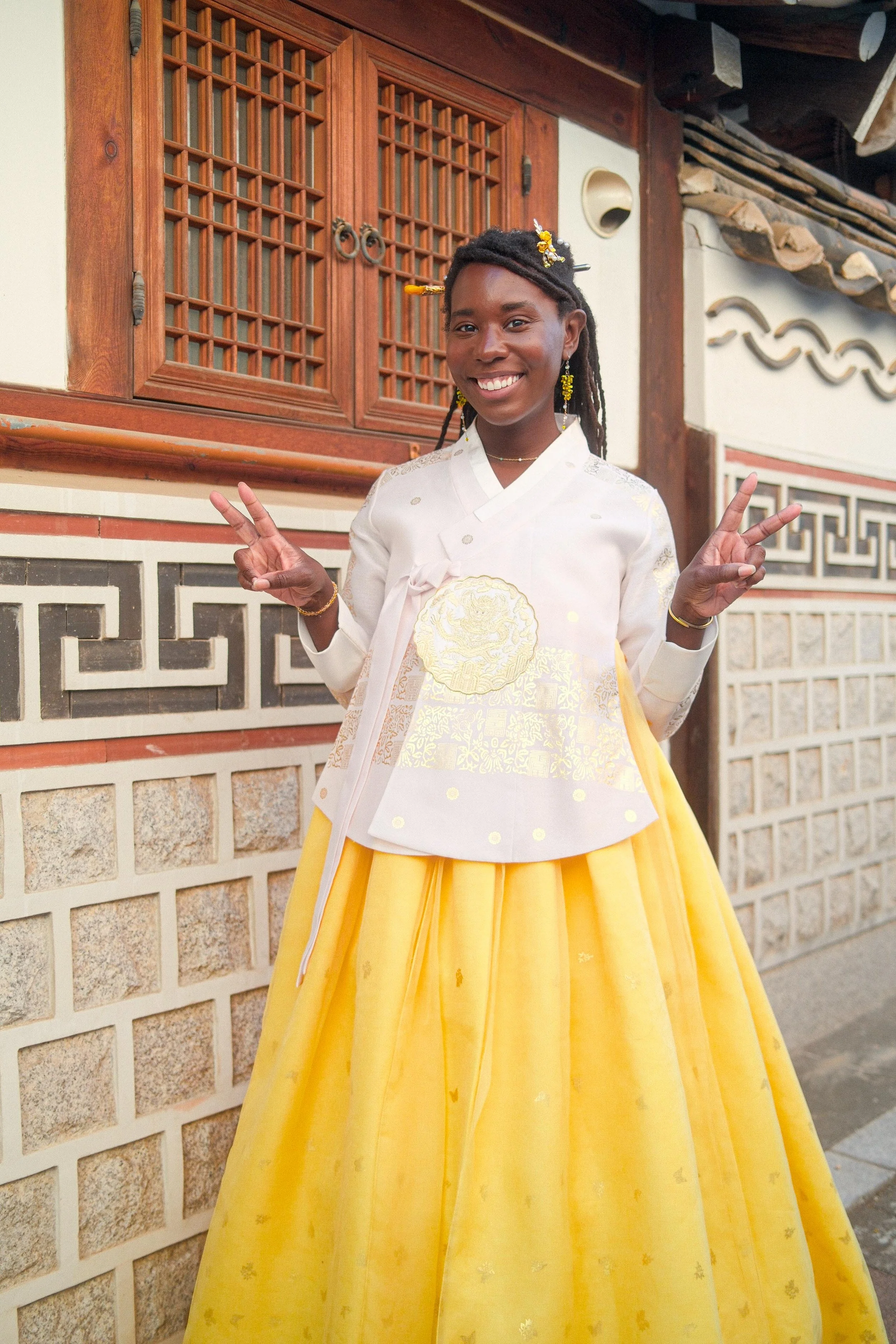 A woman dressed in traditional Korean attire standing in front of a wooden door and brick wall, smiling and making peace signs with both hands.