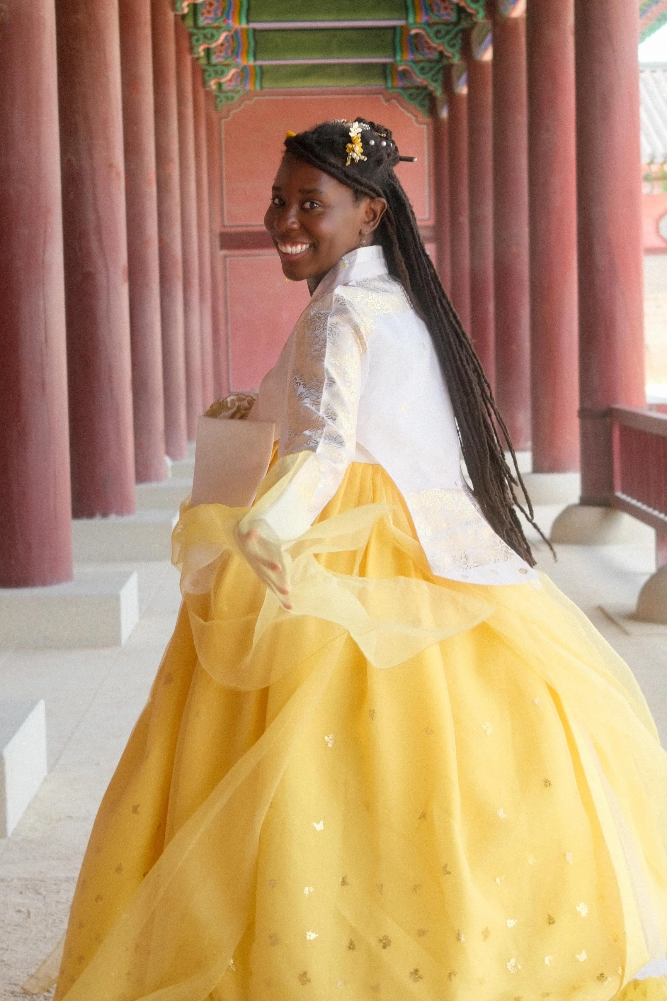 A woman dressed in a traditional Korean hanbok, with a yellow skirt and a white top, smiling and turning her head in an outdoor corridor with pink columns and colorful decorative beams.