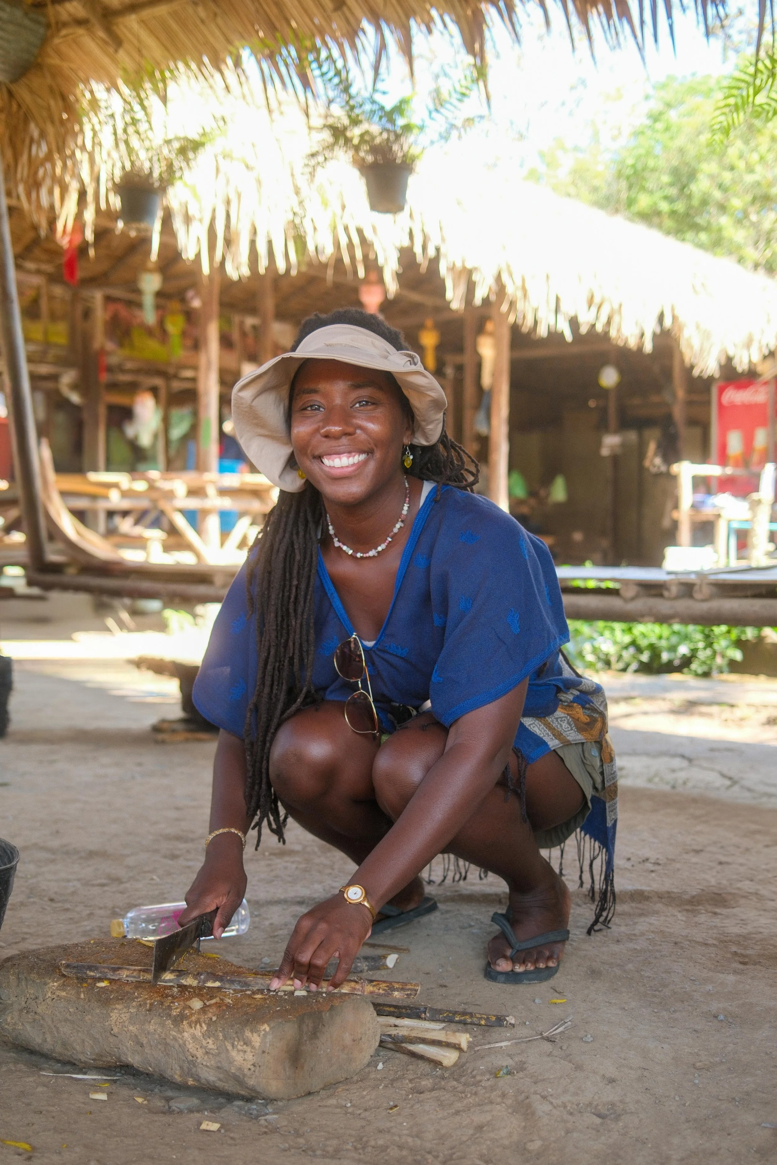 A woman with long dreadlocks and a wide-brimmed hat crouches down and uses a small shovel to work with wood and dirt inside a circular stone structure. She is outdoors, in a rustic setting with a thatched roof hut in the background and a Coca-Cola sign visible.