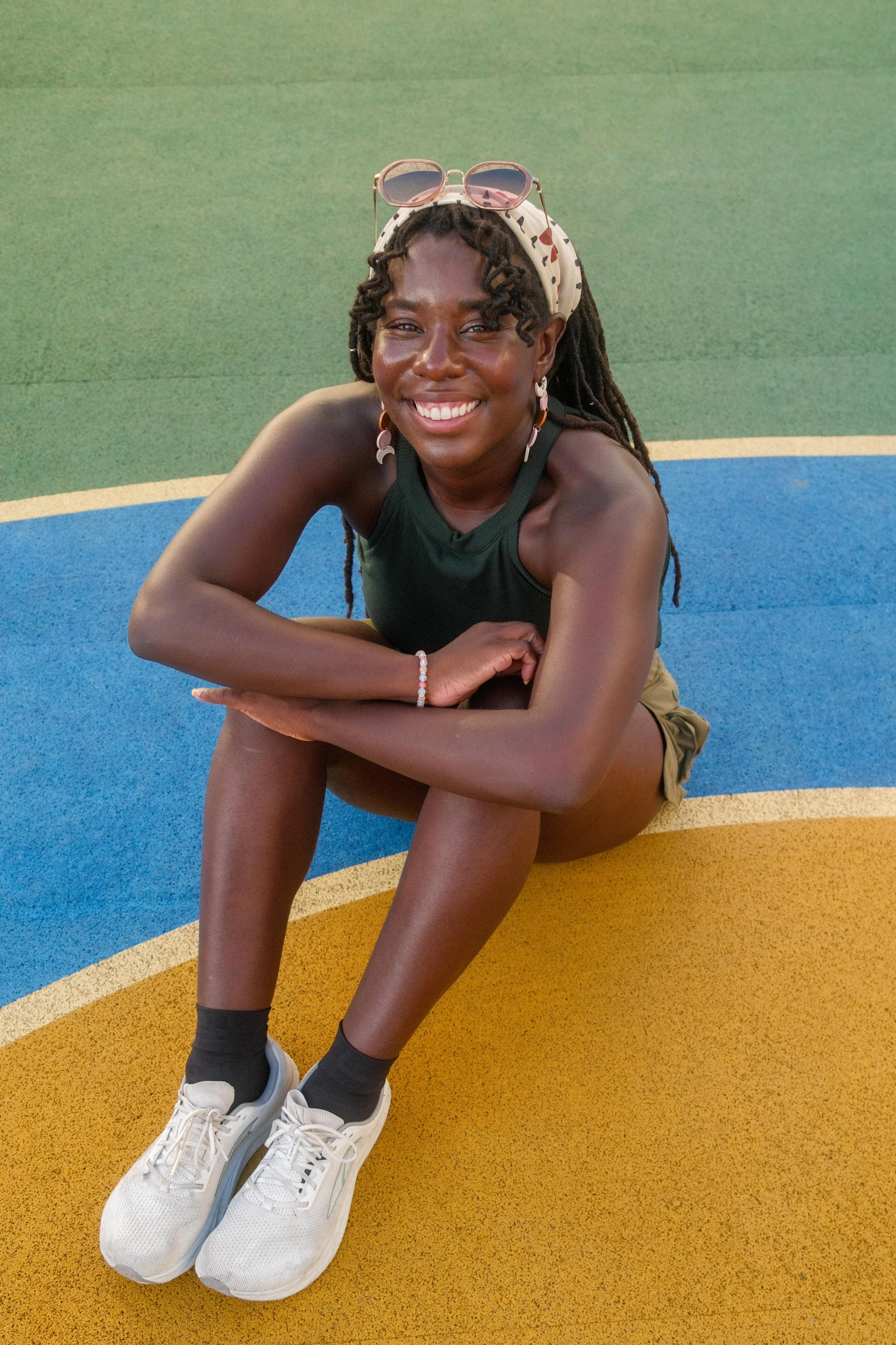 Woman sitting on colorful running track, smiling, wearing a green tank top, tan shorts, black socks, white sneakers, with sunglasses on her head, earrings, and a headscarf.