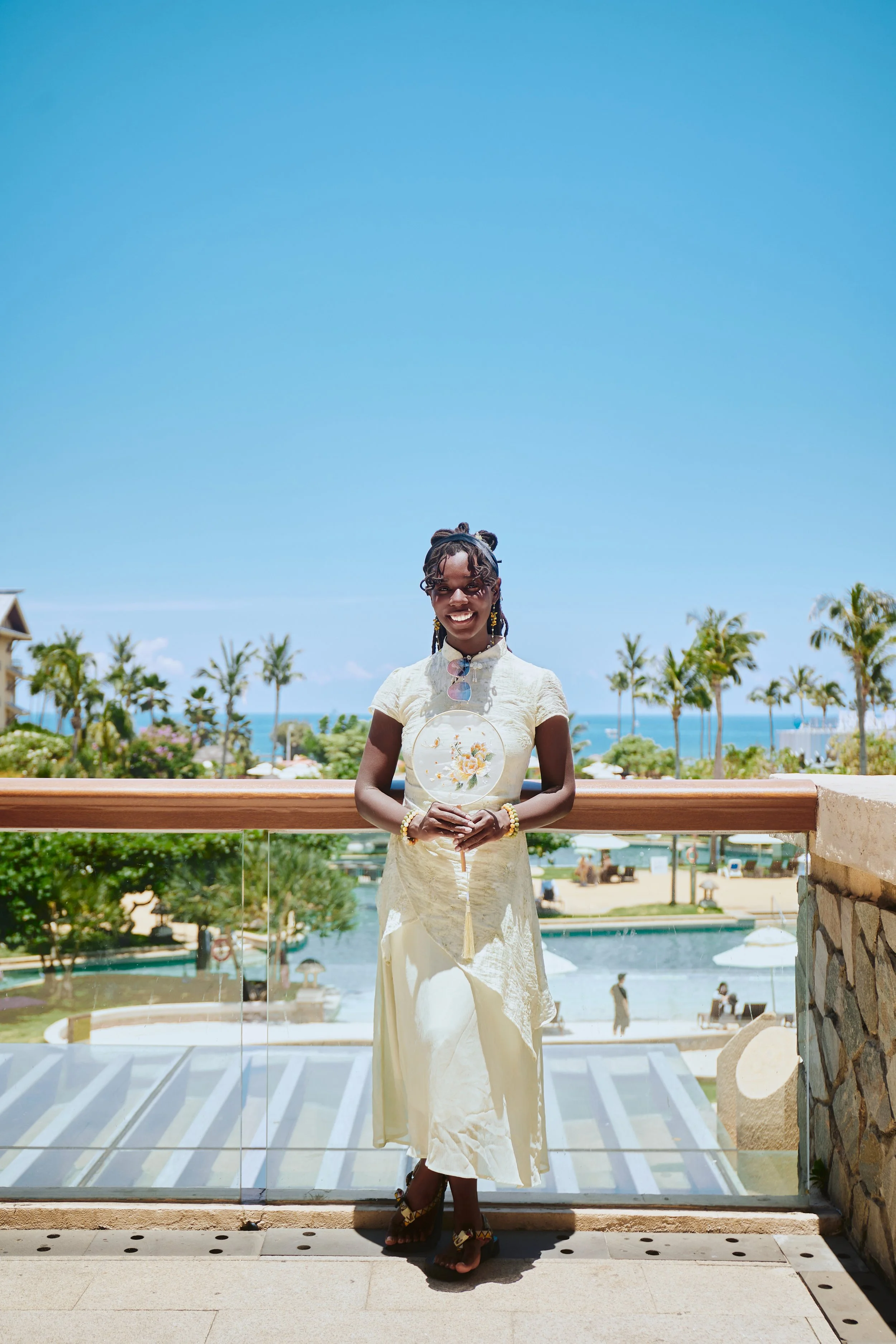 A woman in a cream-colored dress with floral embroidery is standing on a balcony, holding a decorative fan with a floral pattern. She is smiling, wearing jewelry, and has her hair styled in braids with headband. Behind her, there is a tropical landscape with palm trees, a beach, and a clear blue sky.