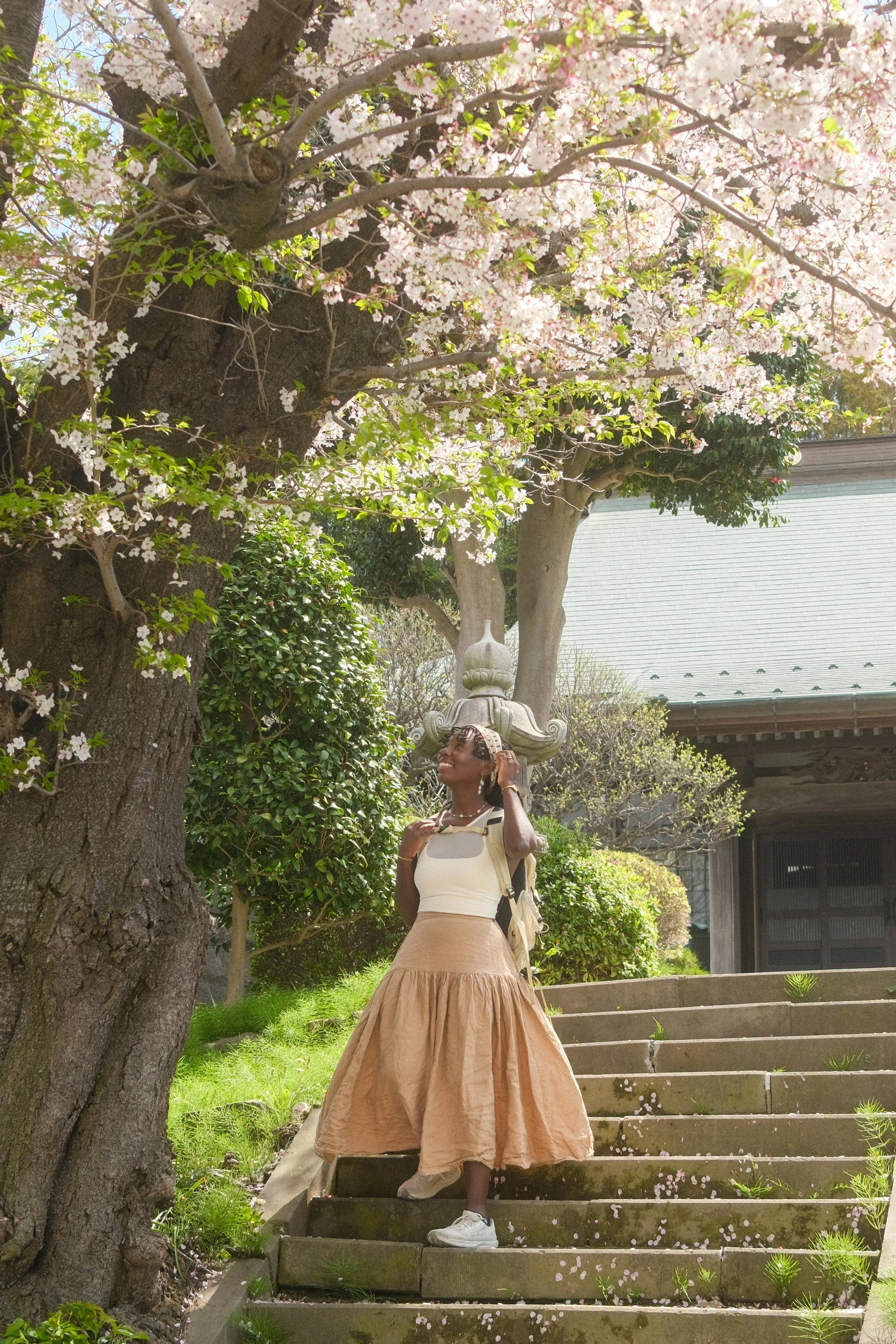 Woman standing on outdoor stairs beneath blooming cherry blossom tree with pink flowers, wearing a white top, beige skirt, white sneakers, and carrying a backpack, smiling and touching her hair.