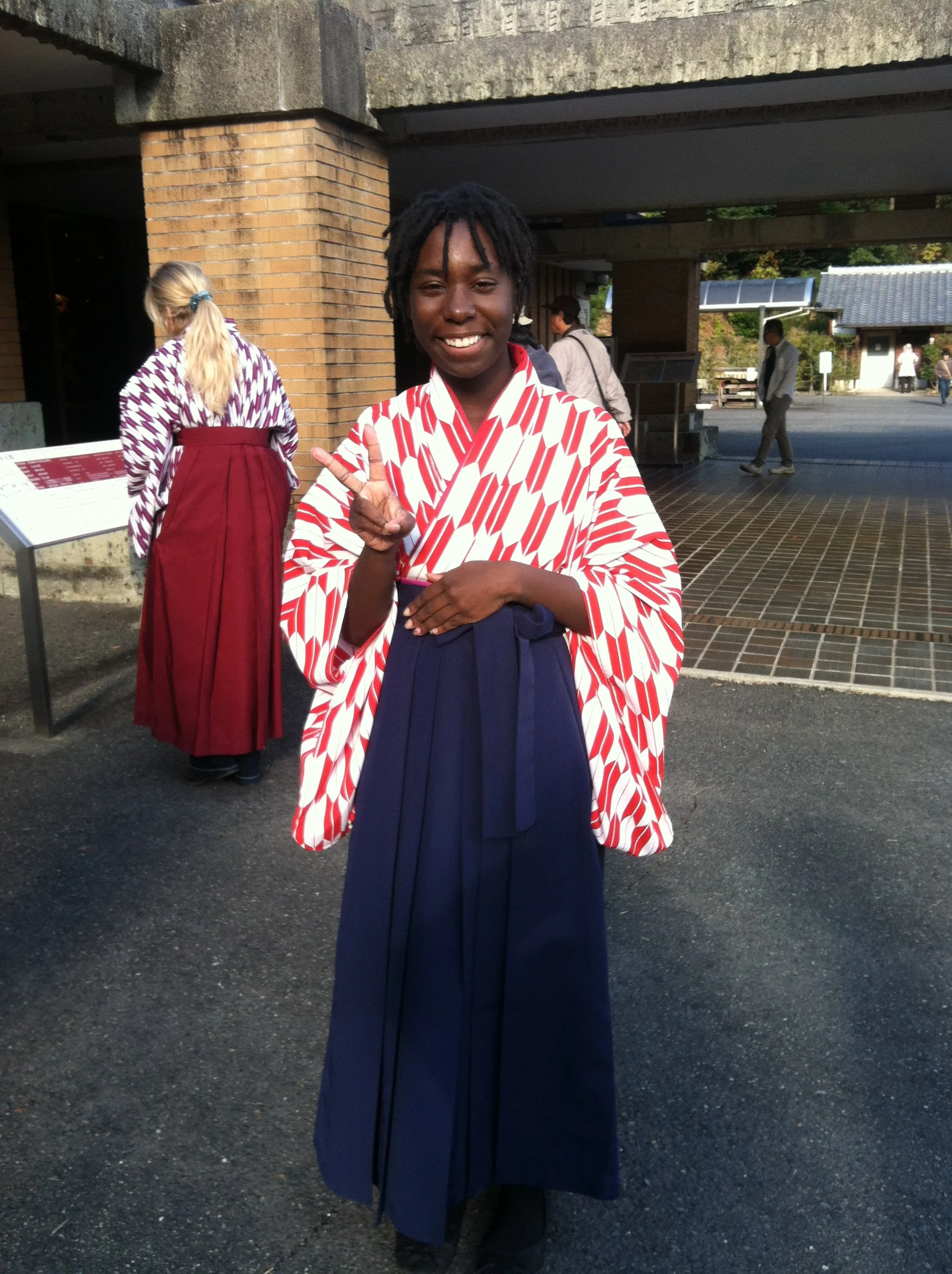 A woman in traditional attire making a peace sign, standing outside near a brick building with other people in the background.
