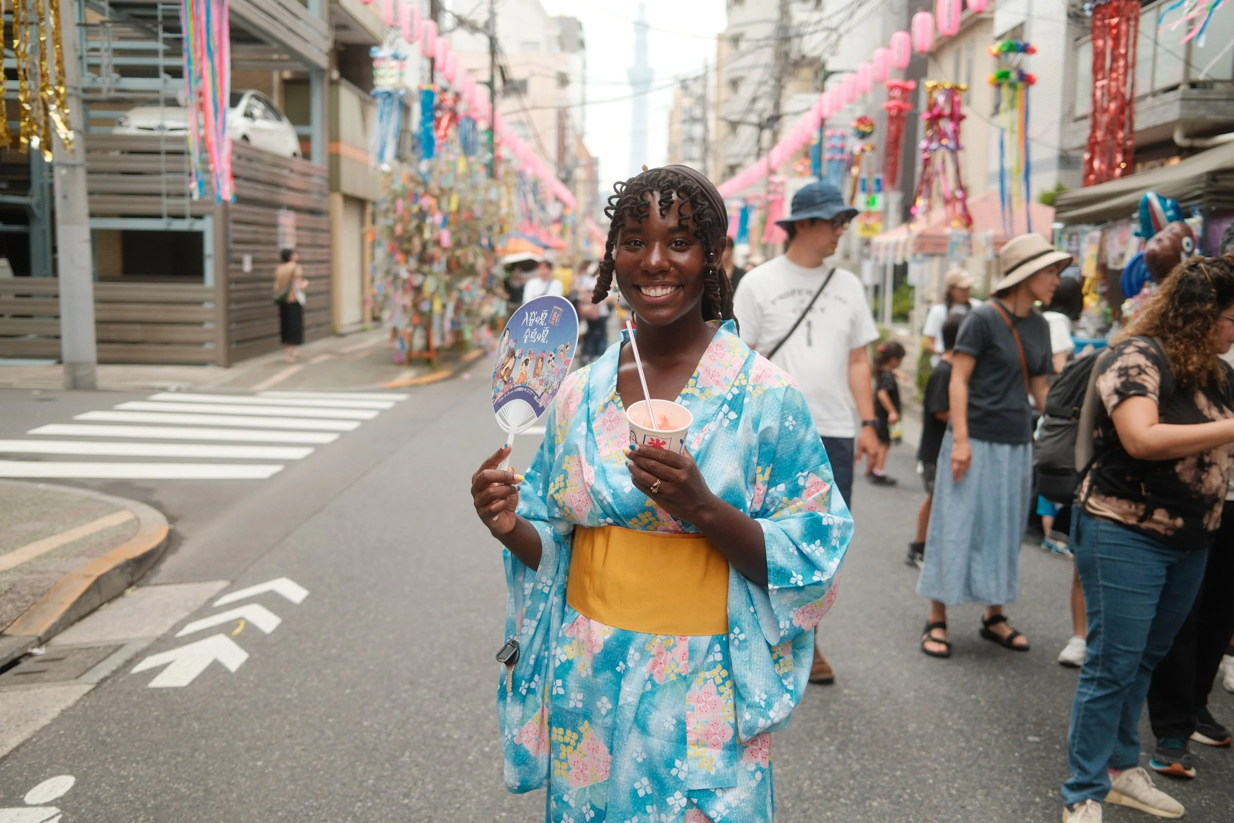 A smiling woman in traditional Japanese attire holding a fan and a cup stands in a street decorated with pink paper lanterns and colorful streamers during a festival, with other festival-goers around her.