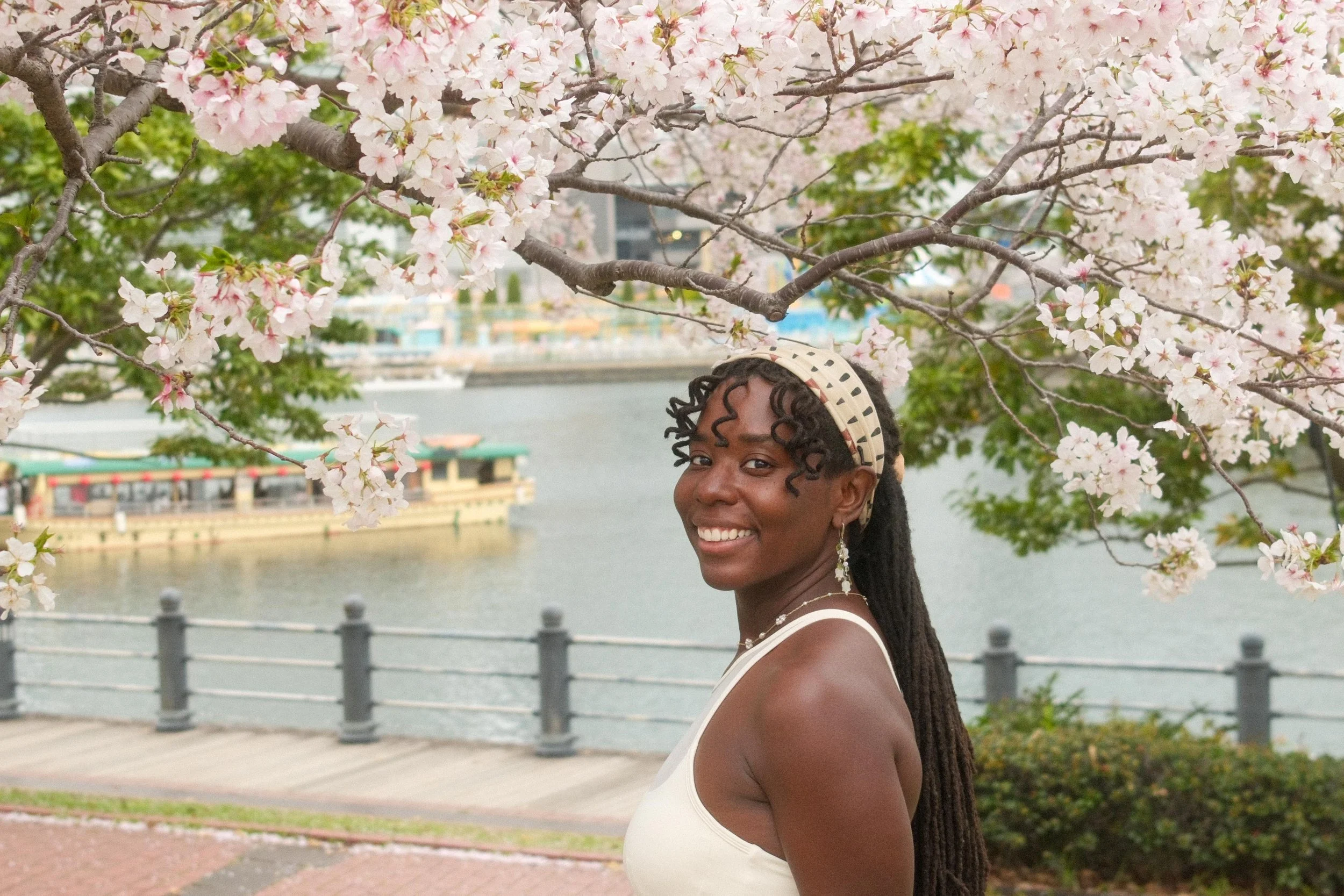 A woman with braided hair and a headband smiling outdoors near a river, with cherry blossoms and trees in the background.