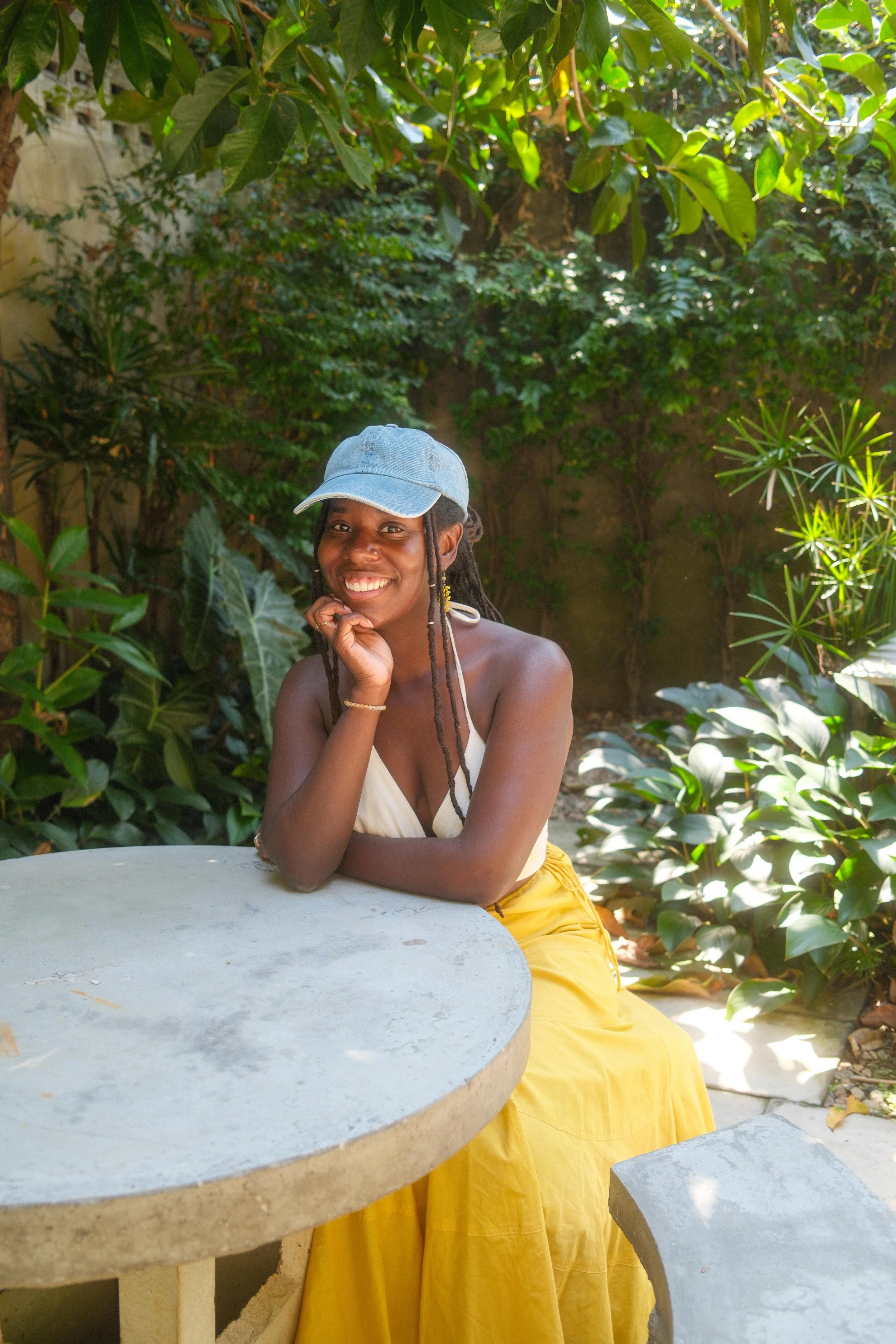 A woman with dark skin, curly hair, and a nose ring, smiling, sitting at an outdoor table surrounded by lush green plants and trees, wearing a light blue cap, white top, and yellow skirt.