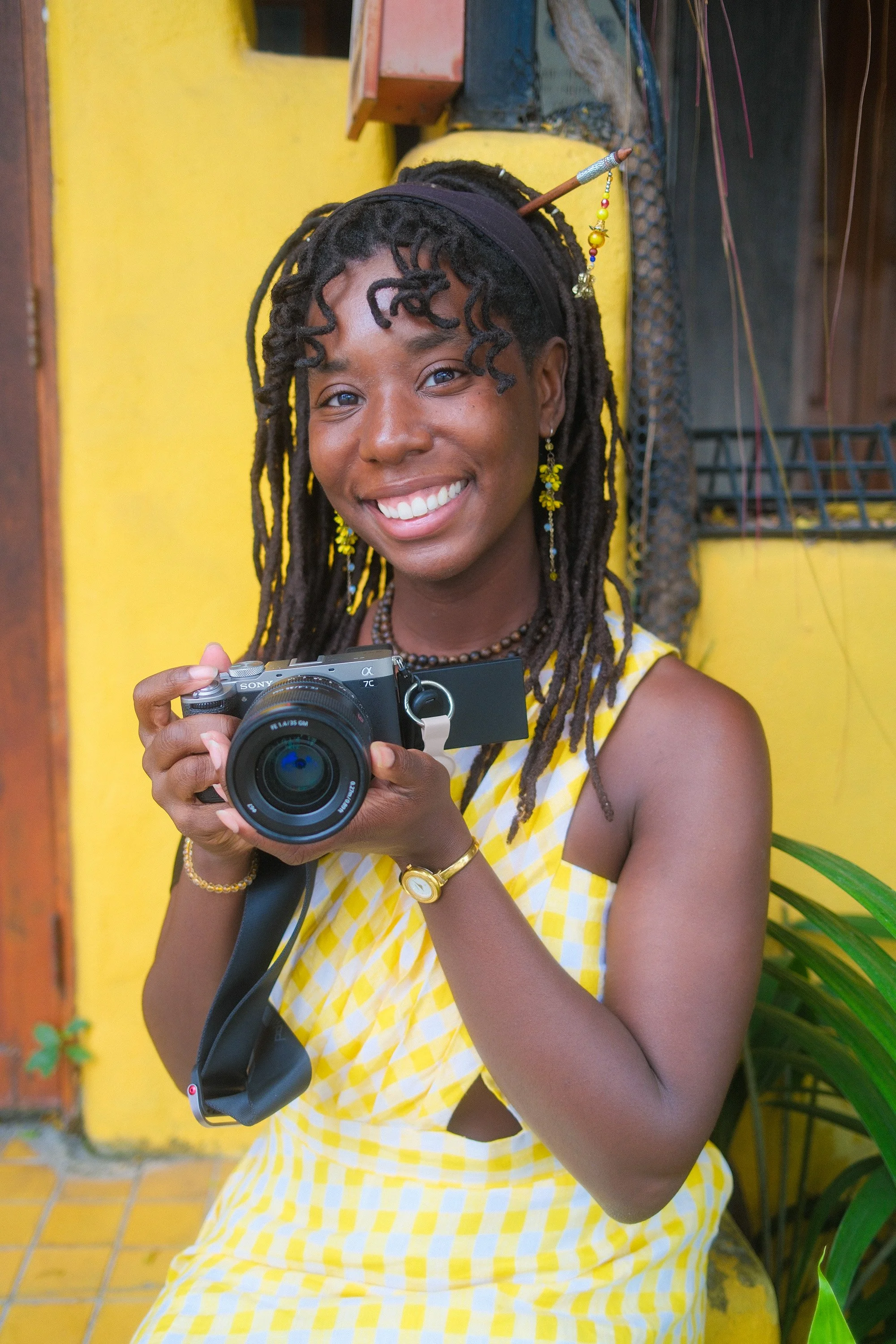 A smiling woman with long dreadlocks dressed in a yellow checked dress, holding a camera, standing outdoors against a yellow wall with greenery.
