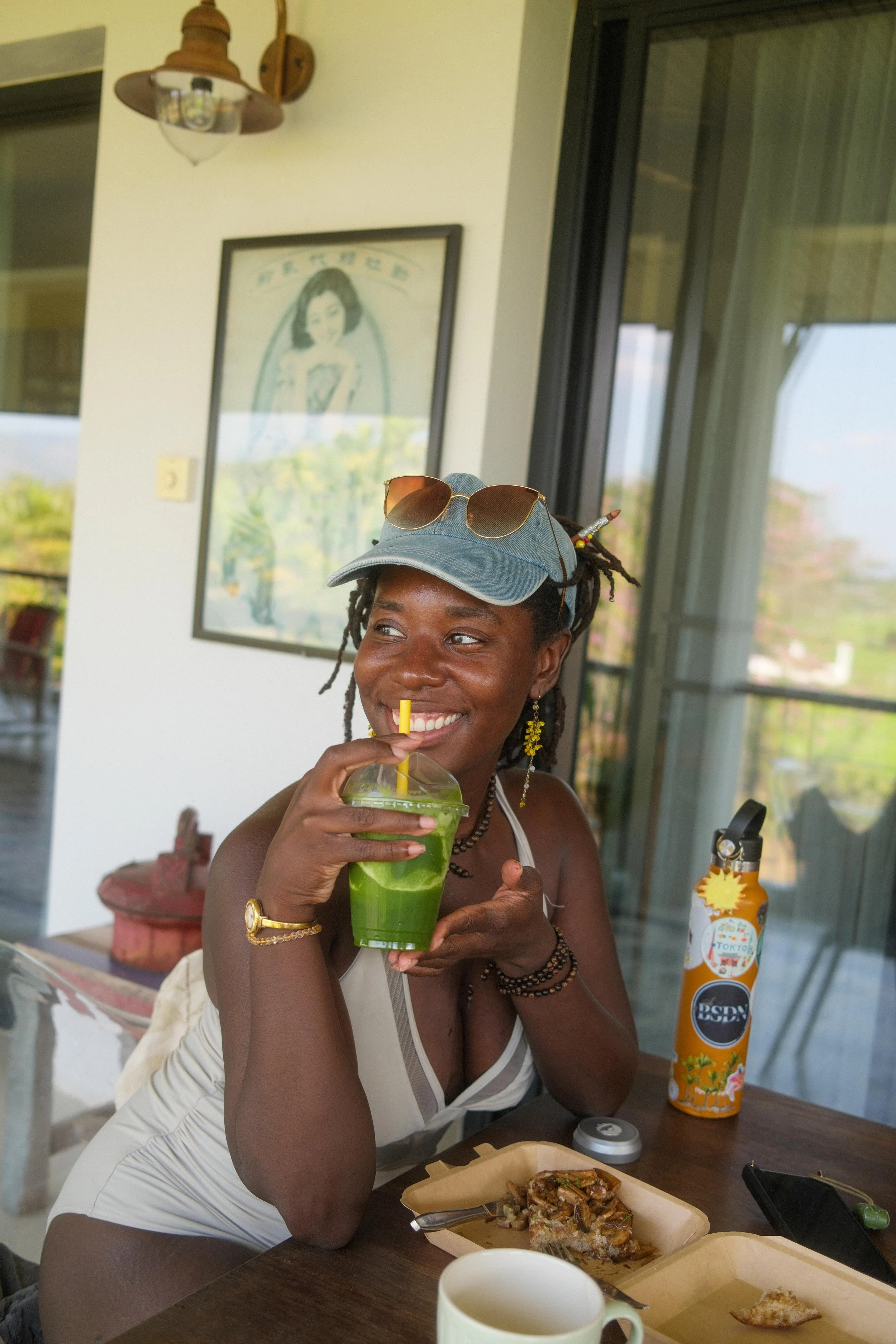 A woman with dark skin, wearing a light-colored tank top, gold jewelry, and sunglasses on a cap, is sitting at a table and enjoying a green drink with a yellow straw. The setting appears to be a casual, indoor-outdoor restaurant or cafe with large glass windows and a framed picture of a woman in the background. There is food on the table, including a slice of cake and a box of food, as well as a bottle of sunscreen or lotion.