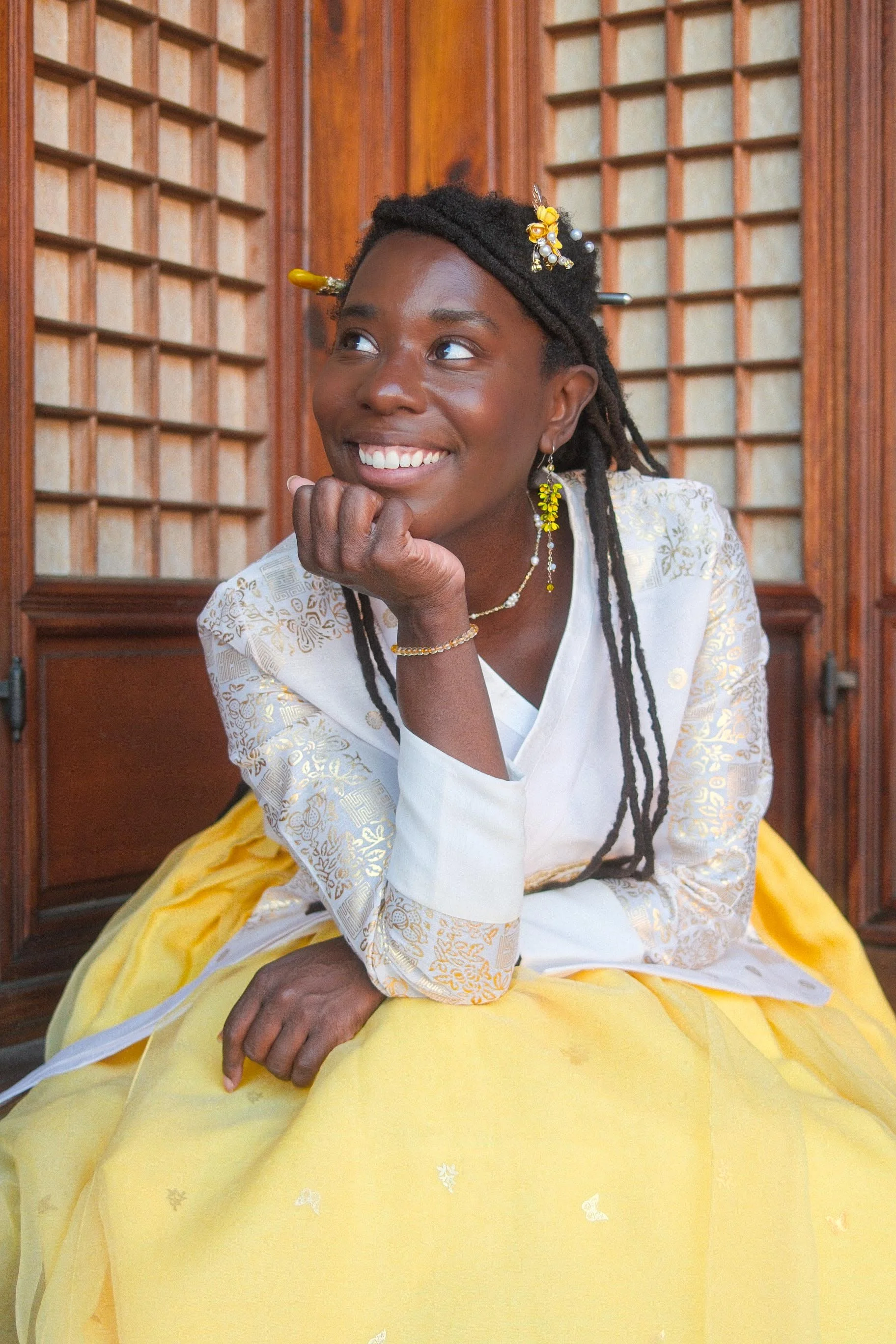 A woman in a yellow and white traditional dress sitting in front of a wooden background, smiling and resting her chin on her hand.