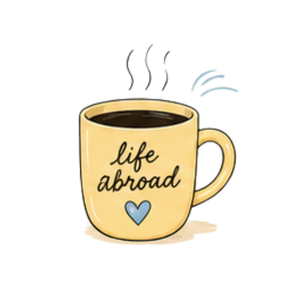 A beige coffee mug with the words 'life abroad' and a blue heart on it, with steam rising from hot coffee inside.