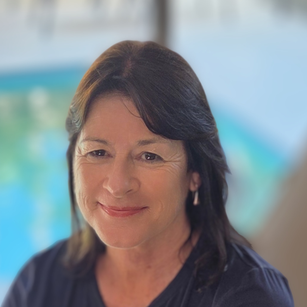 A woman with shoulder-length dark brown hair and a smile, wearing a dark top, with a blurred background that appears to be water.
