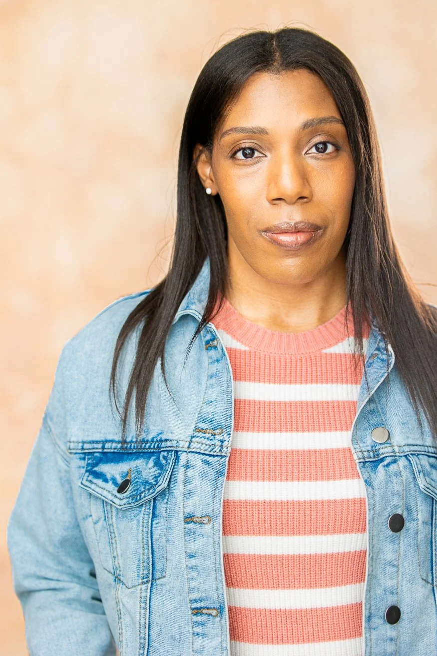 An African American woman with long straight dark-brown hair, wearing a denim jacket over a pink and white striped shirt, standing against a beige background.