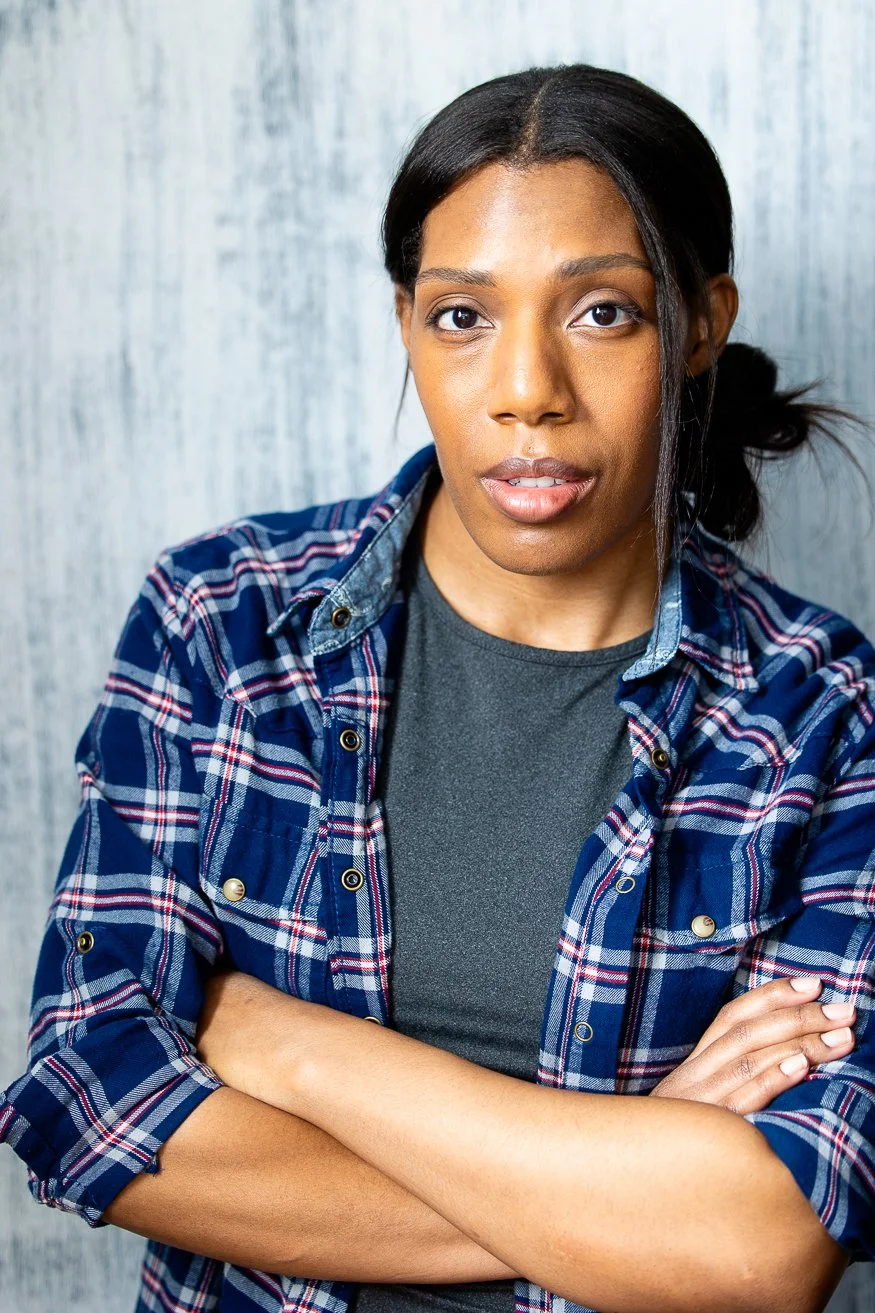 An African American woman with dark-brown hair styled in a side bun, wearing a blue plaid shirt over a gray t-shirt, standing with arms crossed against a light-colored wooden wall.