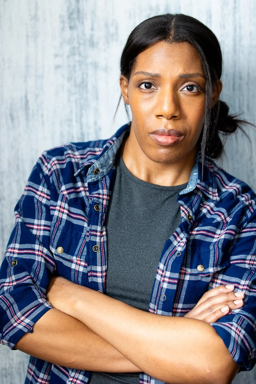An African American woman with dark-brown hair styled in a side bun, wearing a blue plaid shirt over a gray t-shirt, standing with arms crossed against a light-colored wooden wall.