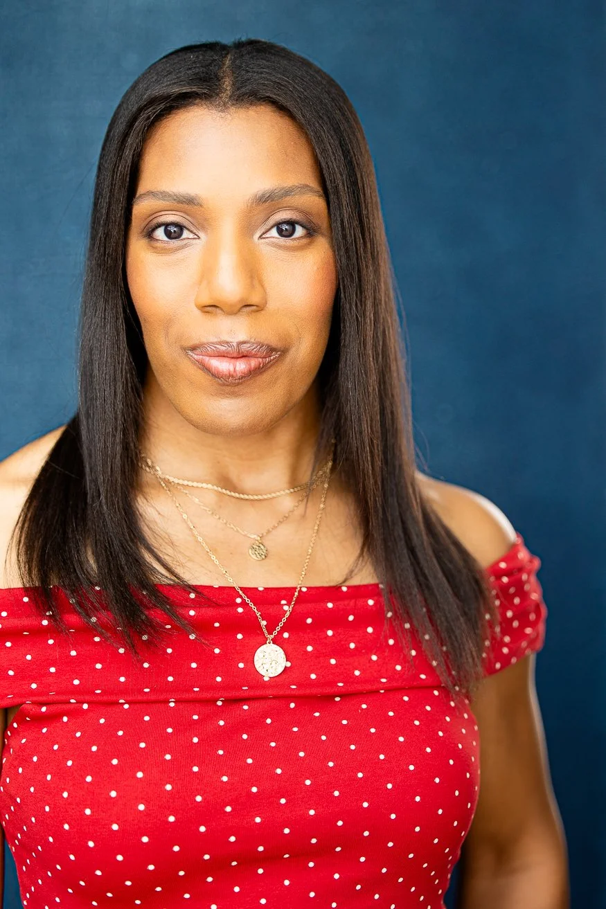 An African American woman with long straight dark-brown hair, wearing a red off-the-shoulder top with white polka dots and gold layered necklace, standing against a dark blue background.
