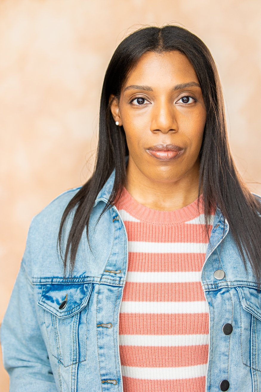 An African American woman with long straight dark-brown hair, wearing a denim jacket over a pink and white striped shirt, standing against a beige background.