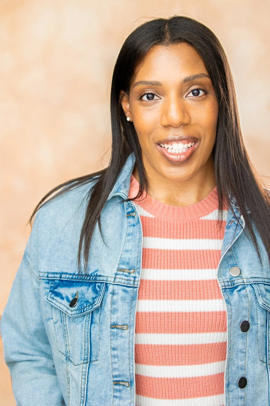 A smiling African American woman with long straight dark-brown hair, wearing a denim jacket over a pink and white striped shirt, standing against a beige background.
