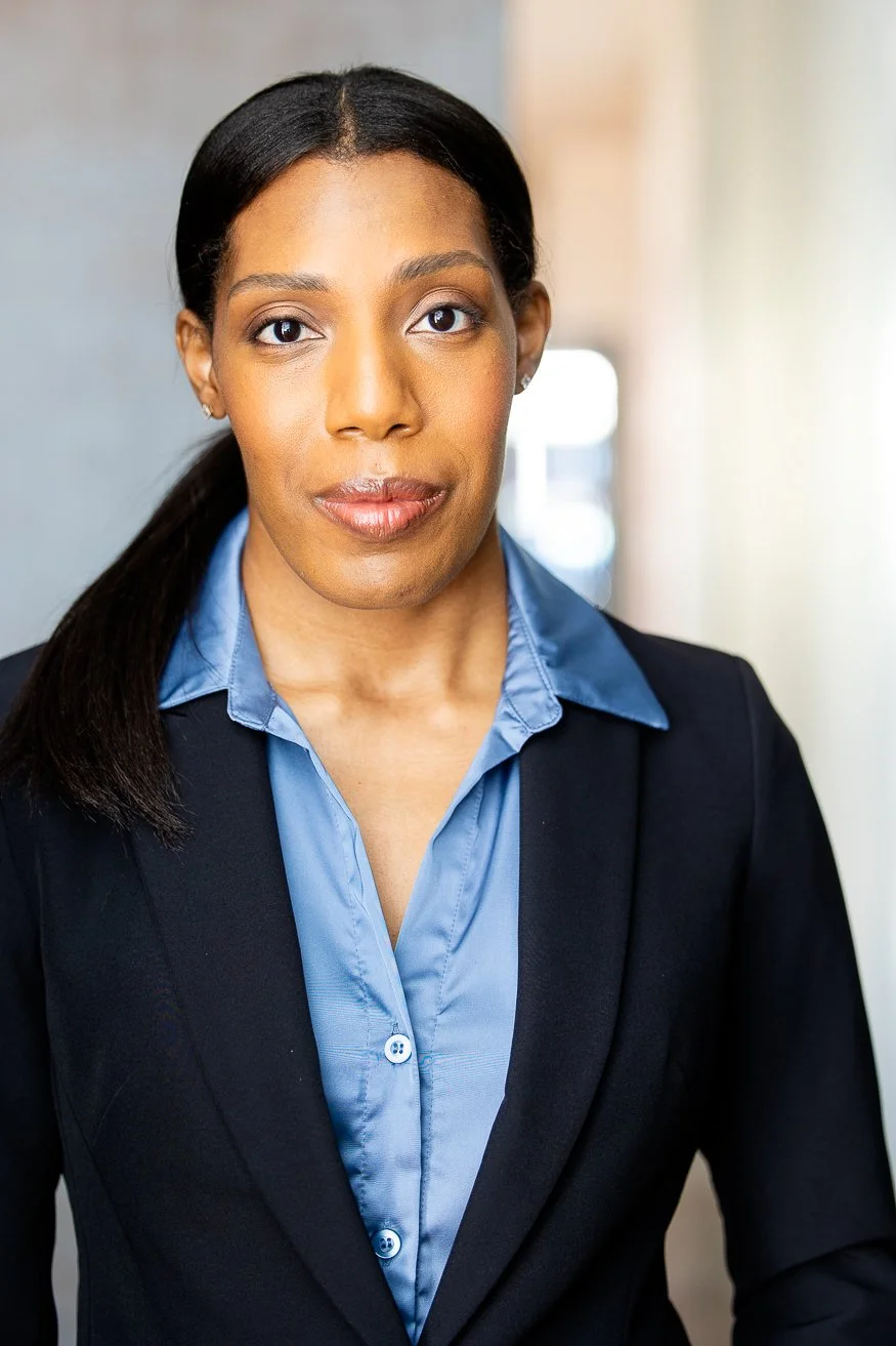 An African American woman with dark-brown hair styled in a ponytail, wearing a black blazer over a blue collared shirt, standing in a well-lit indoor setting.