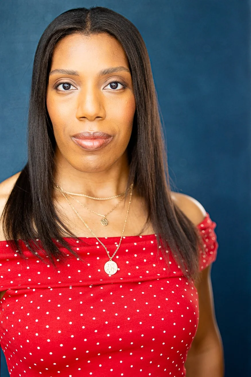 An African American woman with long straight dark-brown hair, wearing a red off-the-shoulder top with white polka dots and gold layered necklace, standing against a dark blue background.
