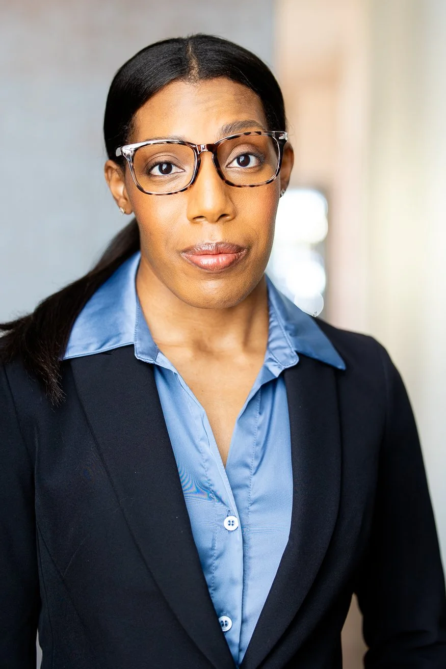 An African American woman with dark-brown hair styled in a ponytail, wearing glasses and a black blazer over a blue collared shirt, standing in a well-lit indoor setting.