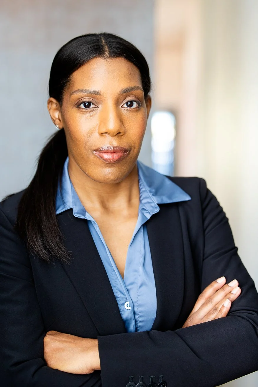 A professional African American woman with dark-brown hair styled in a ponytail, wearing a black blazer over a blue collared shirt, standing with arms crossed in a well-lit indoor setting.